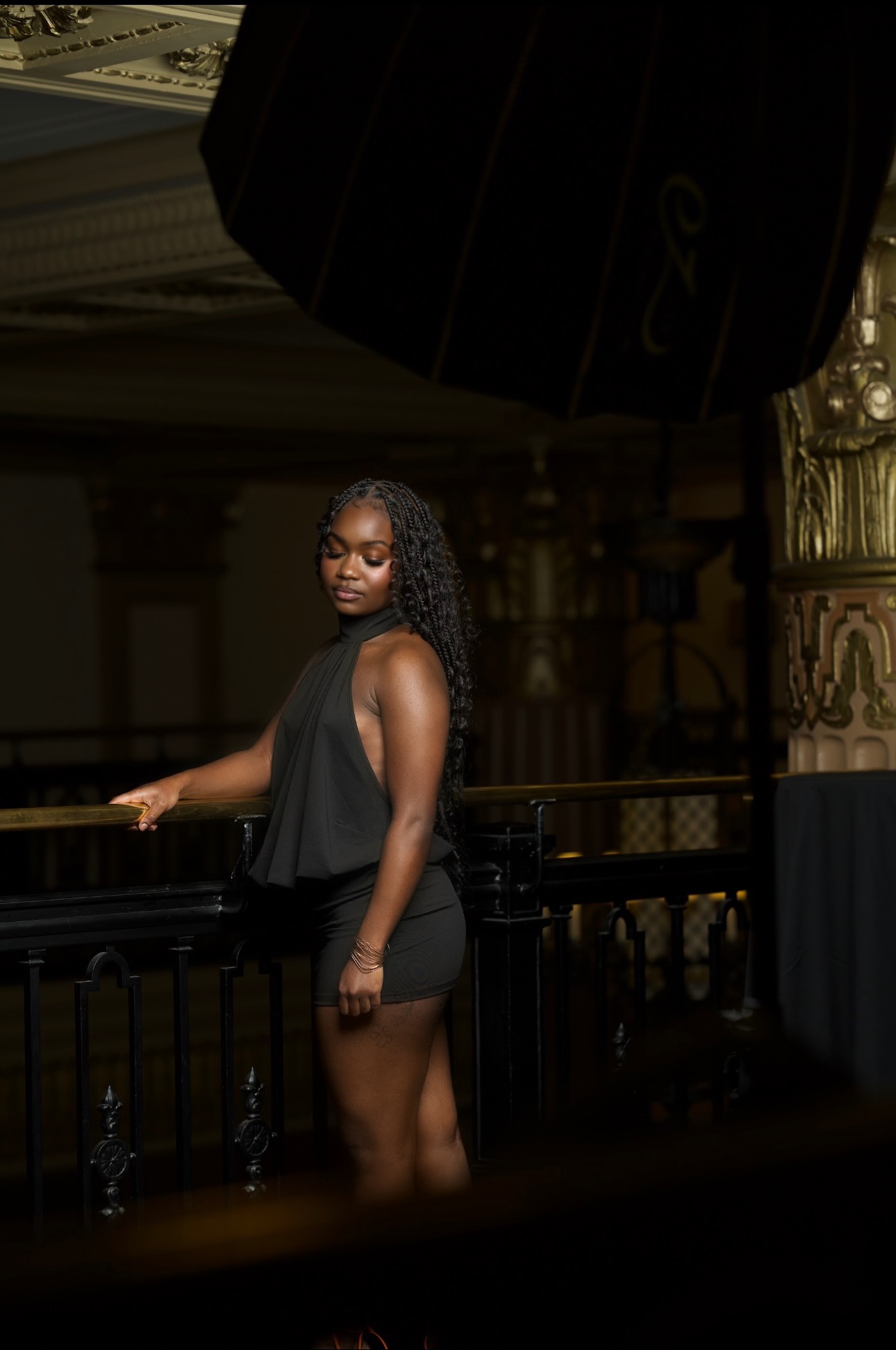 Woman with curly hair in a black dress standing indoors near a railing, with ornate decor in the background.