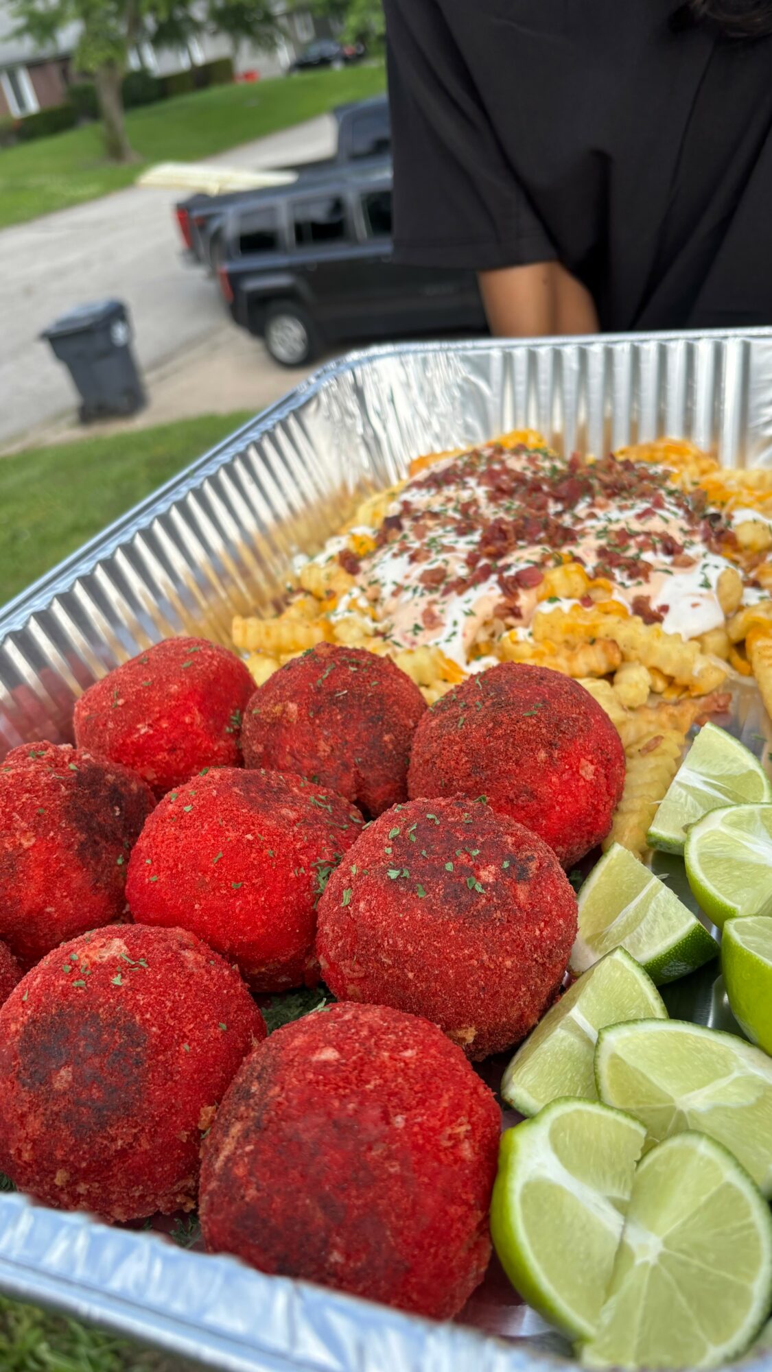 Tray of fried cheese balls with lime wedges and a topping of cheese and sauce, outdoors with a person in black shirt in background.
