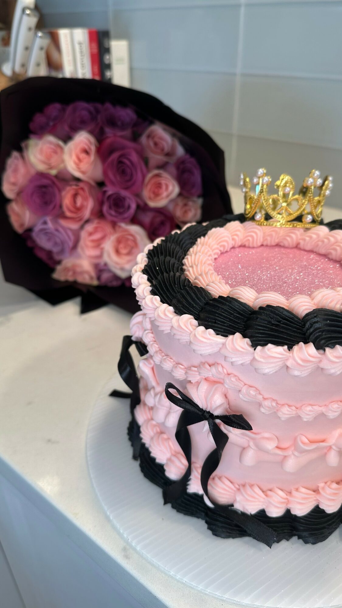 A pink and black layered cake with a gold crown topper, decorated with pink and black icing, and a bouquet of pink and purple roses in the background.