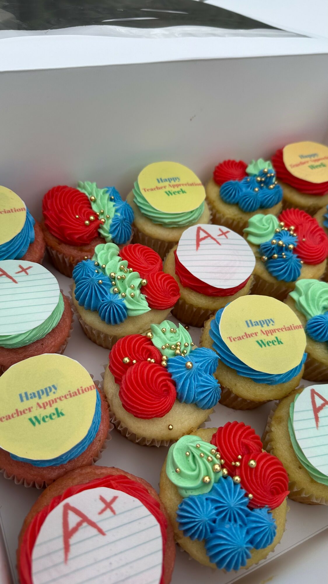 Assorted decorated cupcakes with red, blue, yellow, and green frosting, some with edible toppers, arranged in a box.