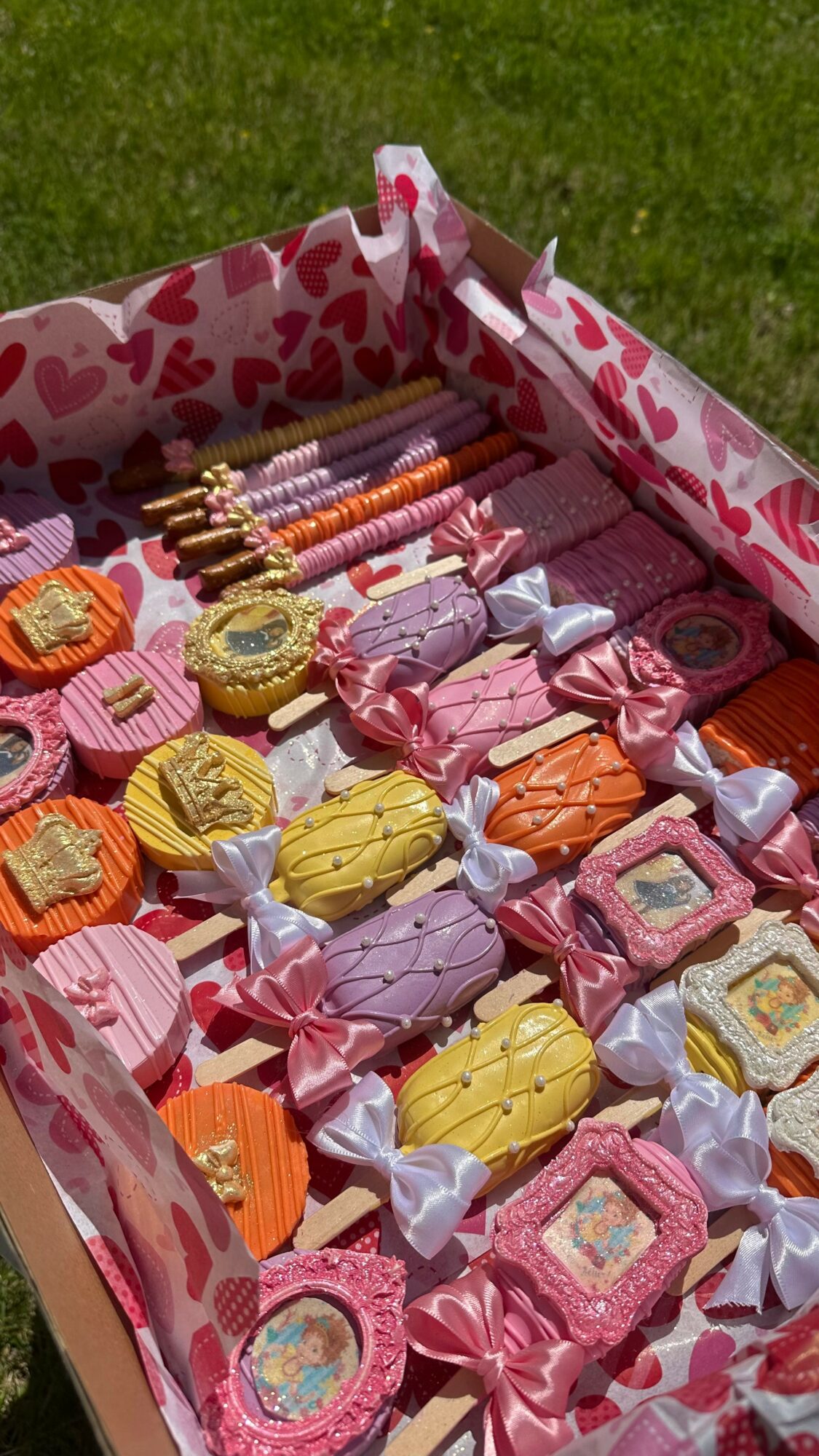 Box filled with colorful wrapped candies and chocolates on grass, with pink and white heart-patterned lining.
