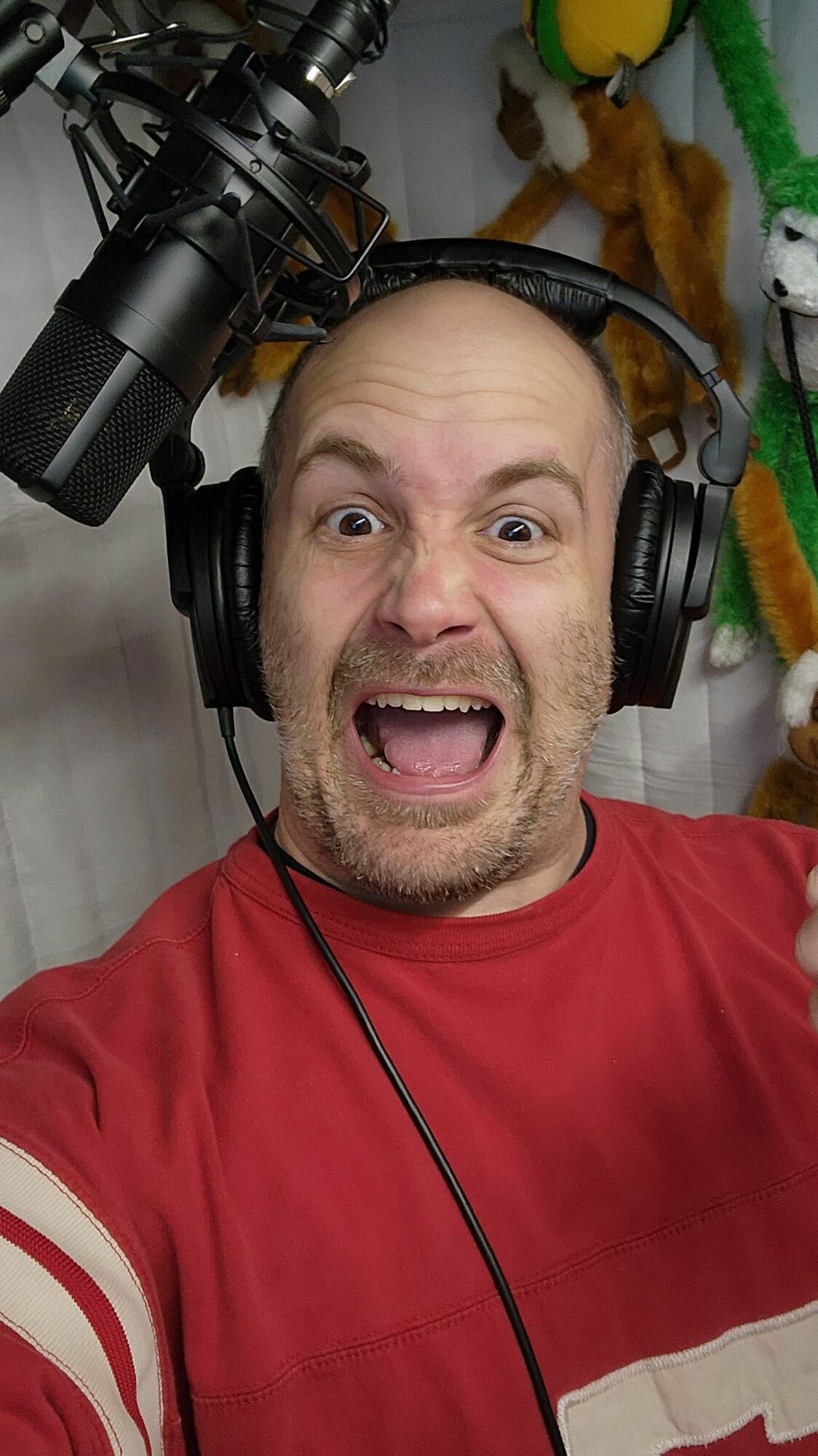Man with headphones speaking into a microphone, wearing a red shirt, in a recording studio.