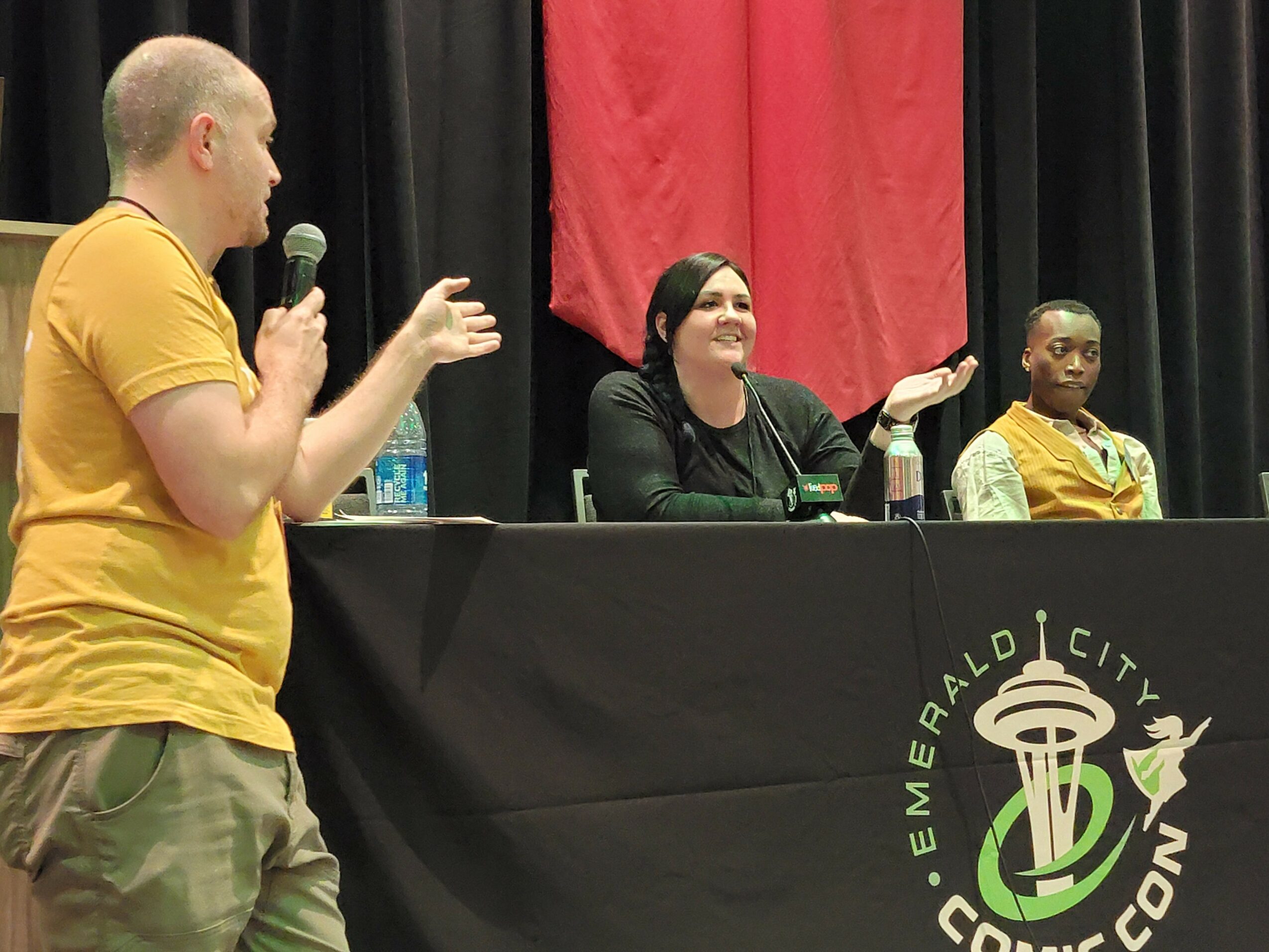 Three people seated at a panel table on stage, with a woman speaking and a person standing with a microphone, black curtains behind, and a red curtain in the background.