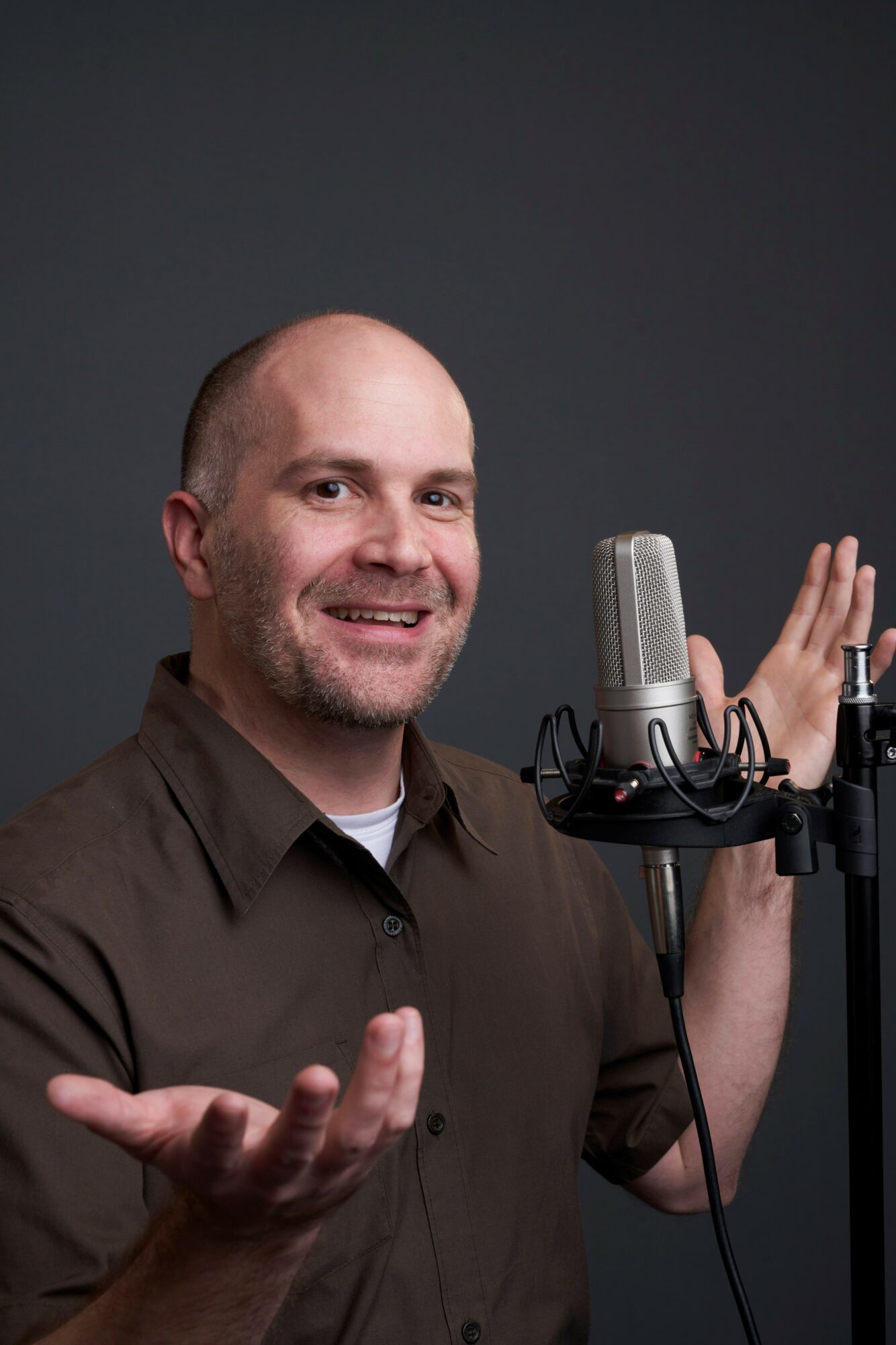 Man with a beard and bald head speaking into a microphone, gesturing with his hands, against a dark background.