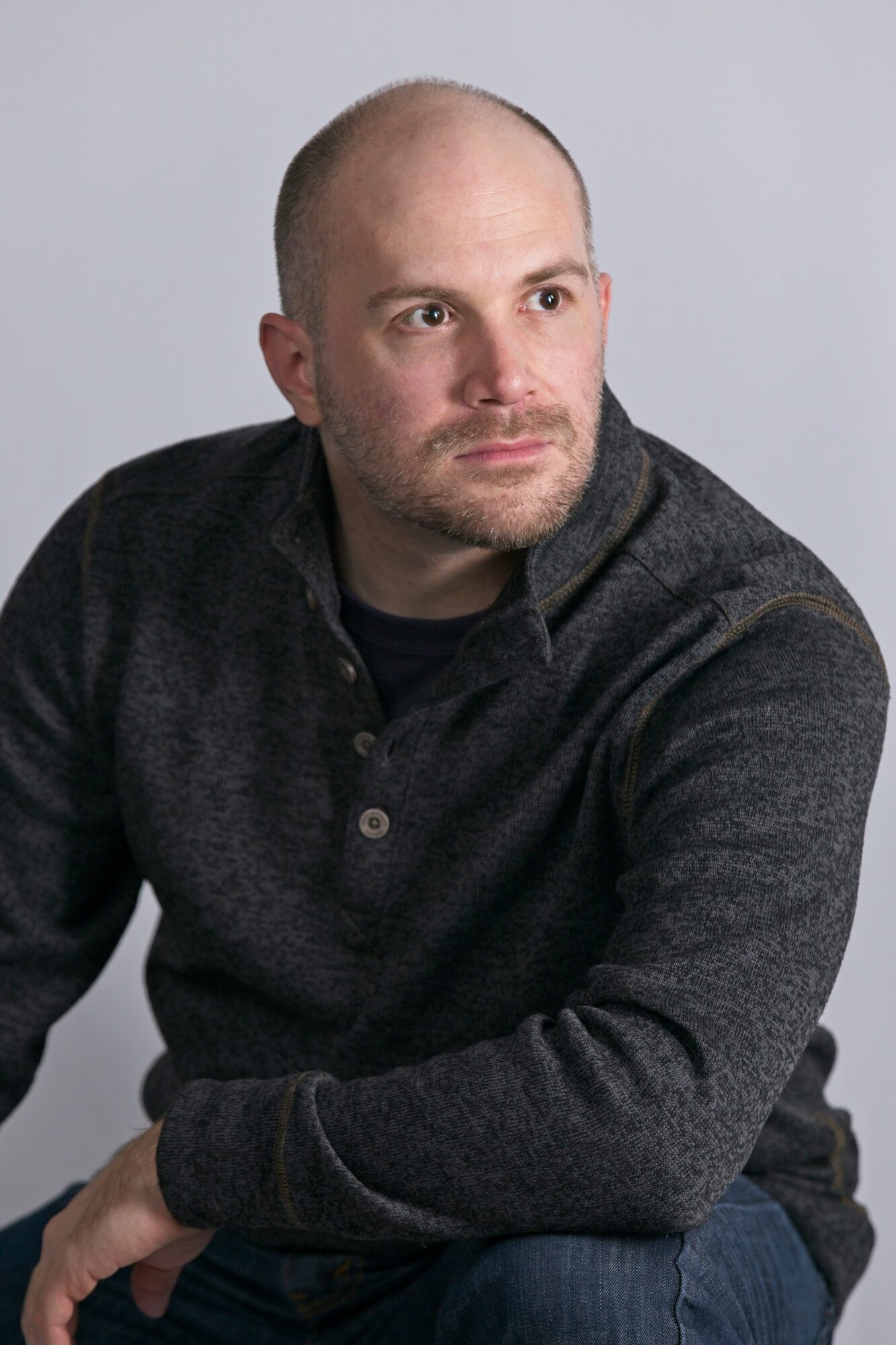 Man with a beard and shaved head wearing a dark gray sweater, looking to the side against a light gray background.