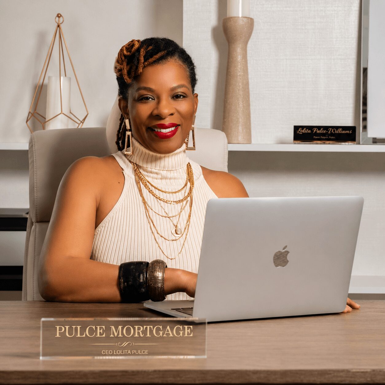 Woman with braided hair and red lipstick sitting at desk with laptop, wearing jewelry and sleeveless top.