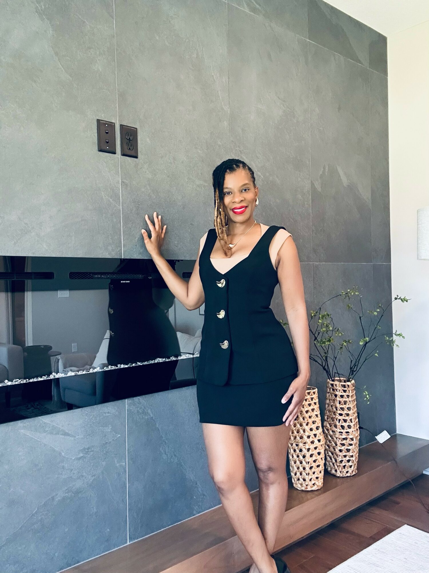 Woman in black dress with buttons, standing indoors near a gray wall and decorative vases, smiling and posing with one hand on the wall.