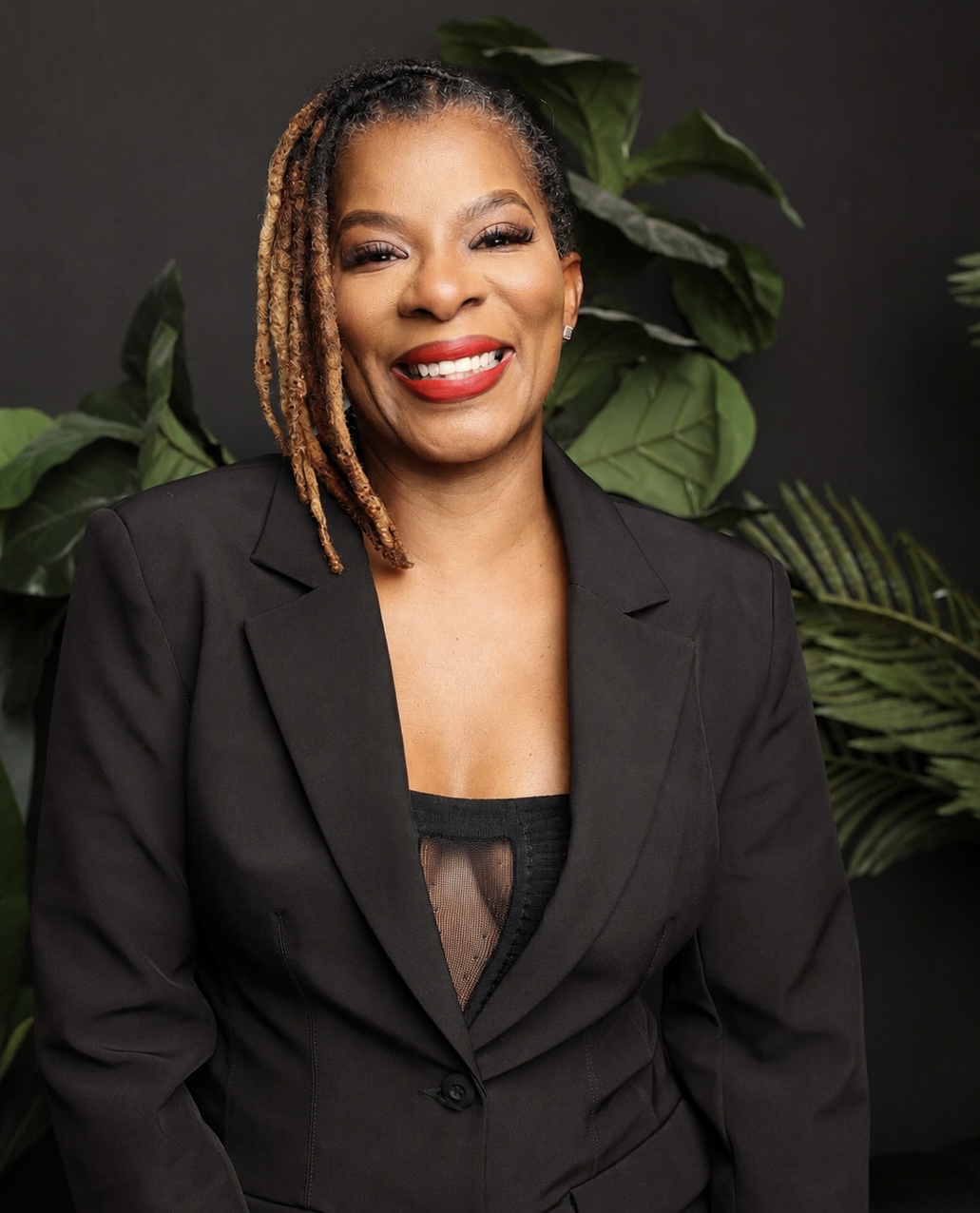 Smiling woman with dreadlocks wearing a black blazer, standing in front of green plants, dark background.
