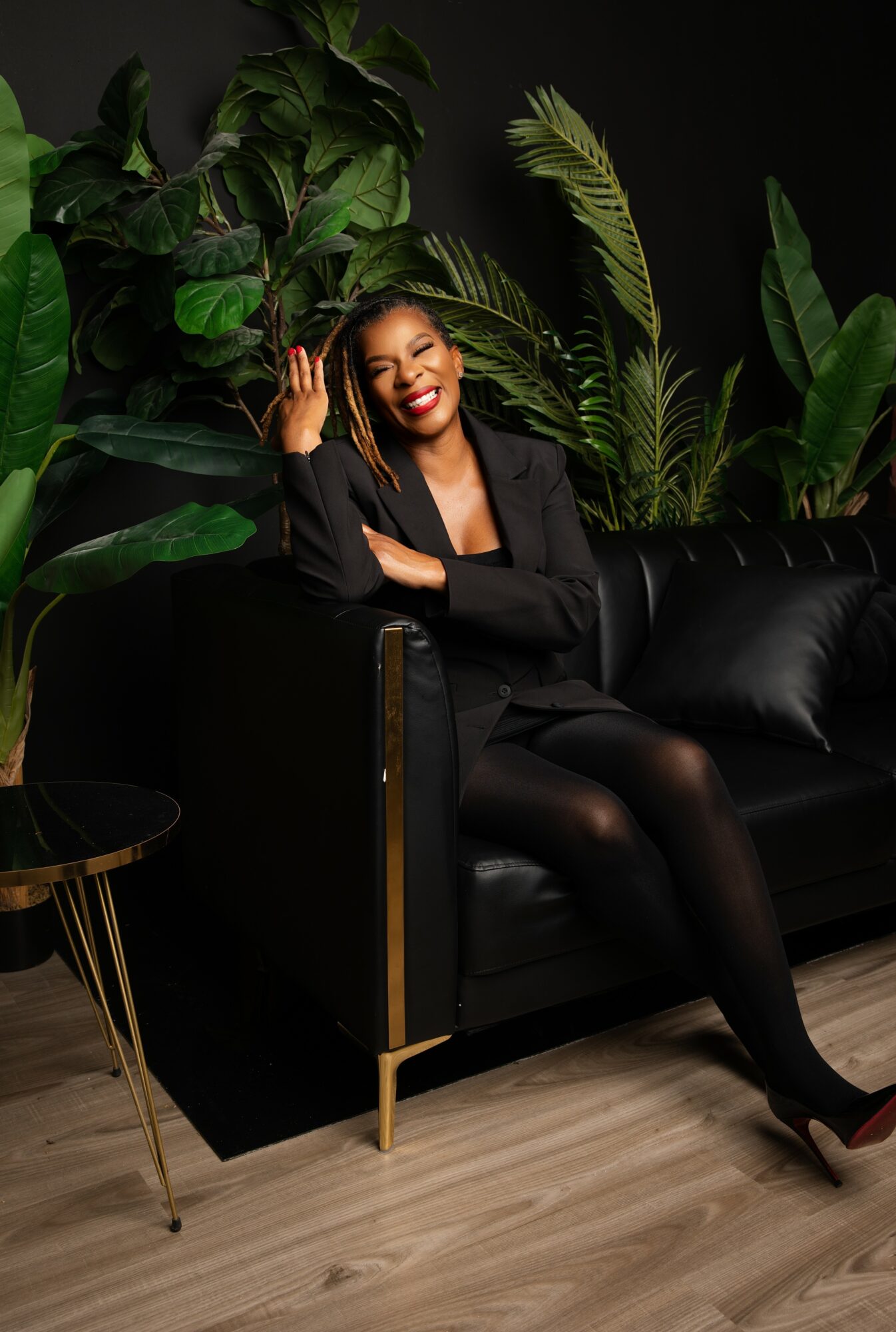 Woman sitting on a black sofa, smiling, surrounded by large green plants, wearing a black blazer and tights, waving.