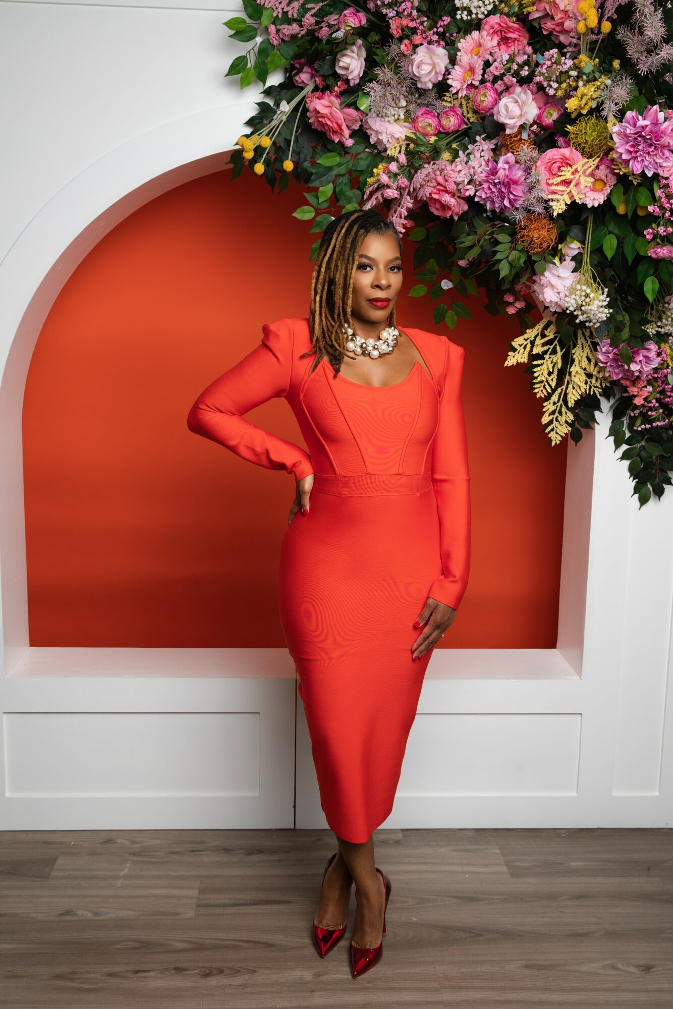 Woman in red dress and heels standing in front of floral display with orange background, posing with hand on hip.