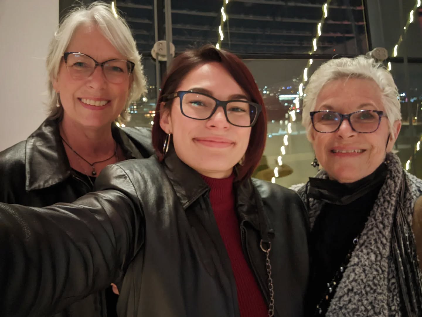 Three women smiling indoors, wearing glasses and dark jackets, with string lights in the background.