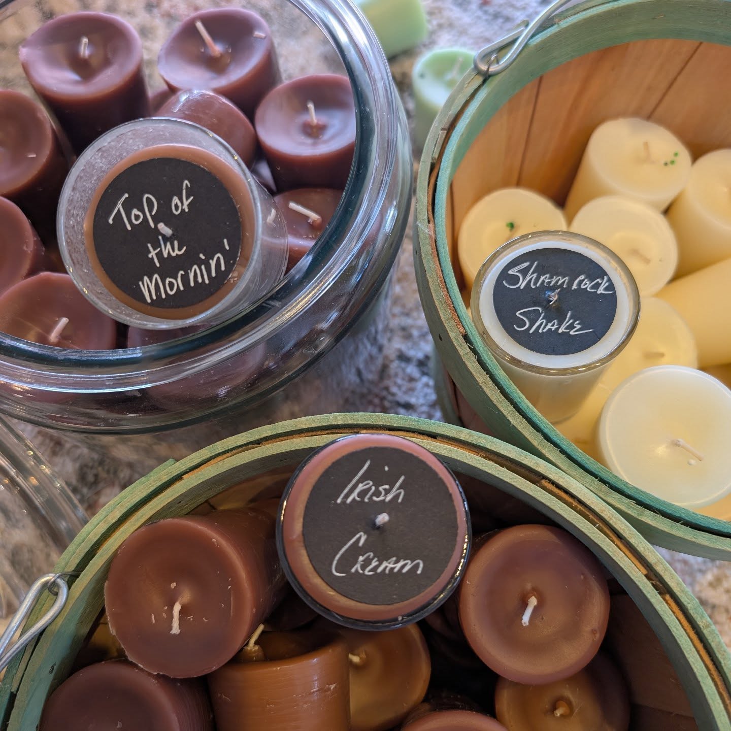 Three baskets of candles with chalkboard labels: top left labeled 'Top of the Morning,' top right labeled 'Shamrock Shake,' bottom labeled 'Irish Cream,' with candles in various colors and shapes.