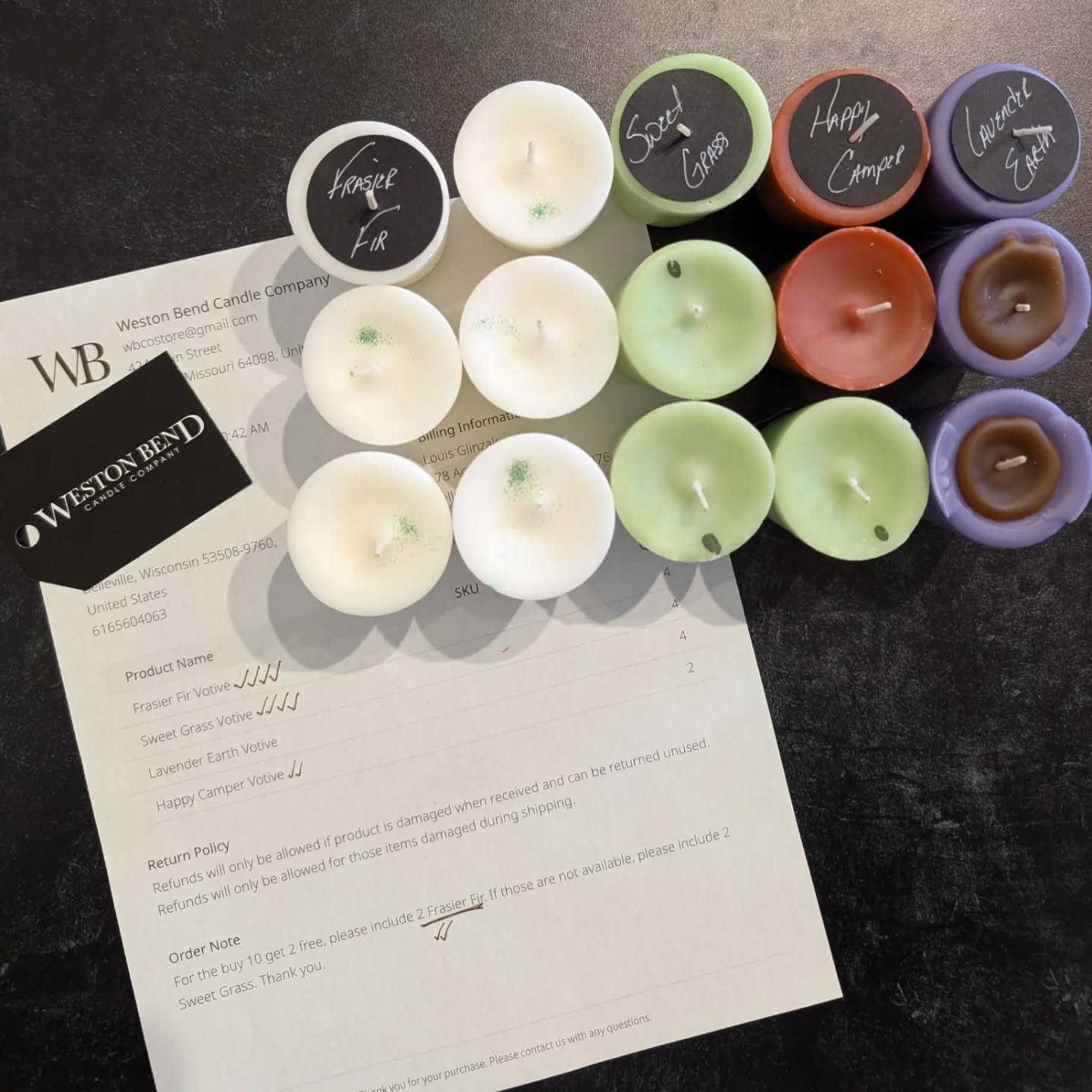 Sixteen candles in various colors arranged on a dark surface with a document and business card nearby.