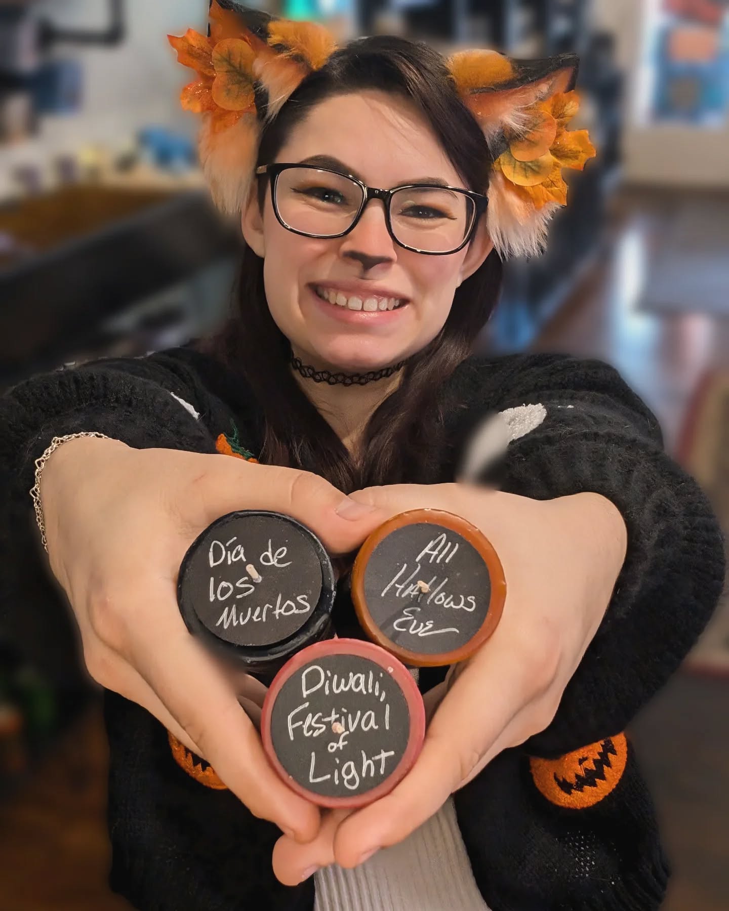 Person wearing animal ears and glasses, holding three heart-shaped signs with Spanish text, smiling, in an indoor setting.