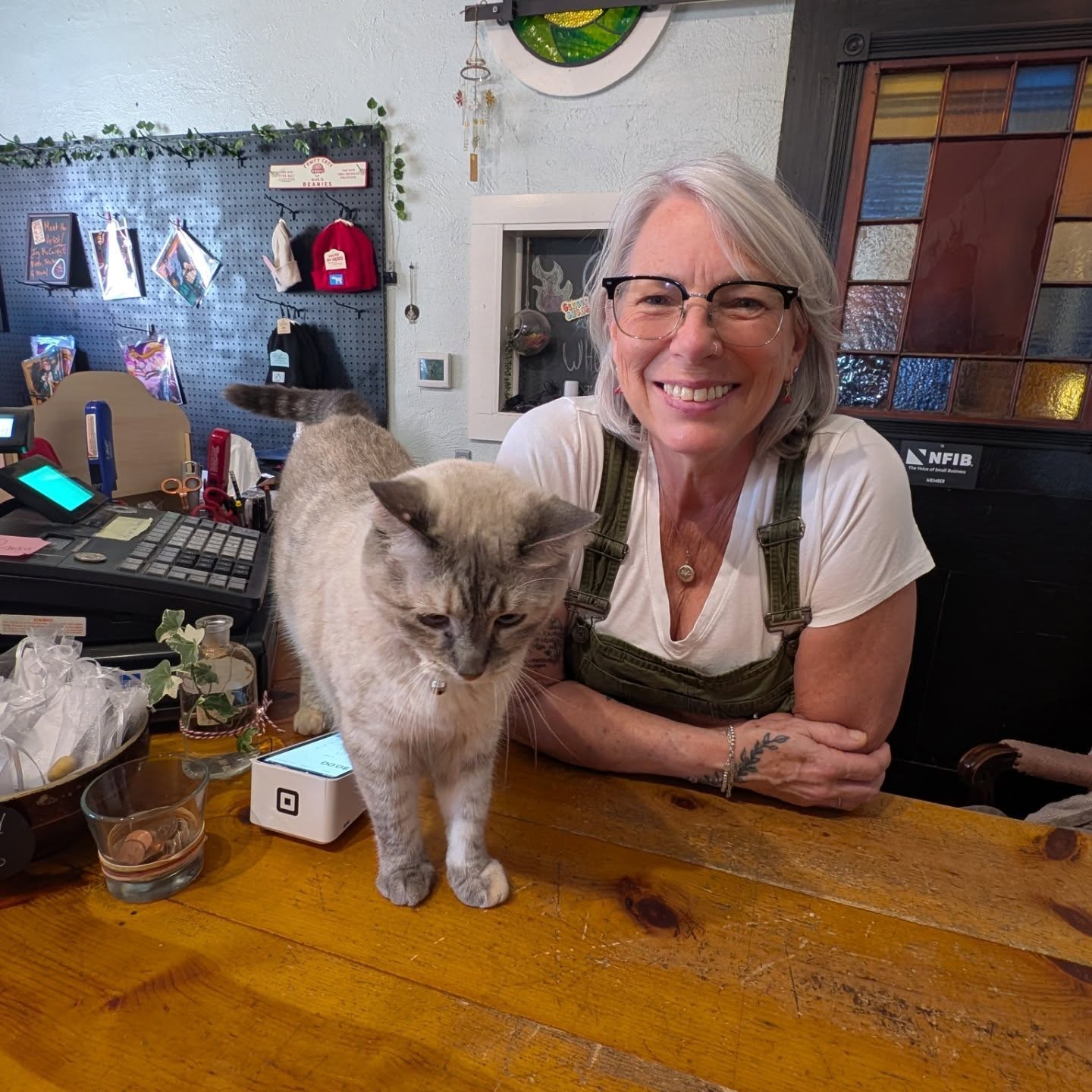 Smiling woman with glasses and gray hair leaning on a wooden counter, with a gray cat nearby, in a cozy room.