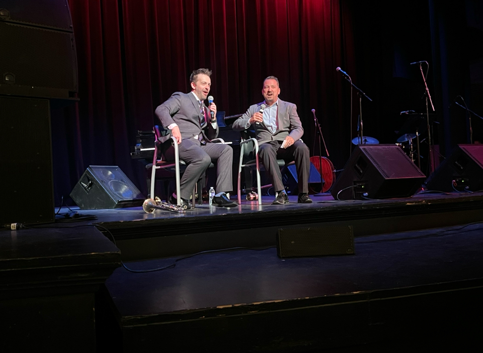 Two men sit on stage with microphones, engaged in conversation, with musical instruments and equipment behind them.