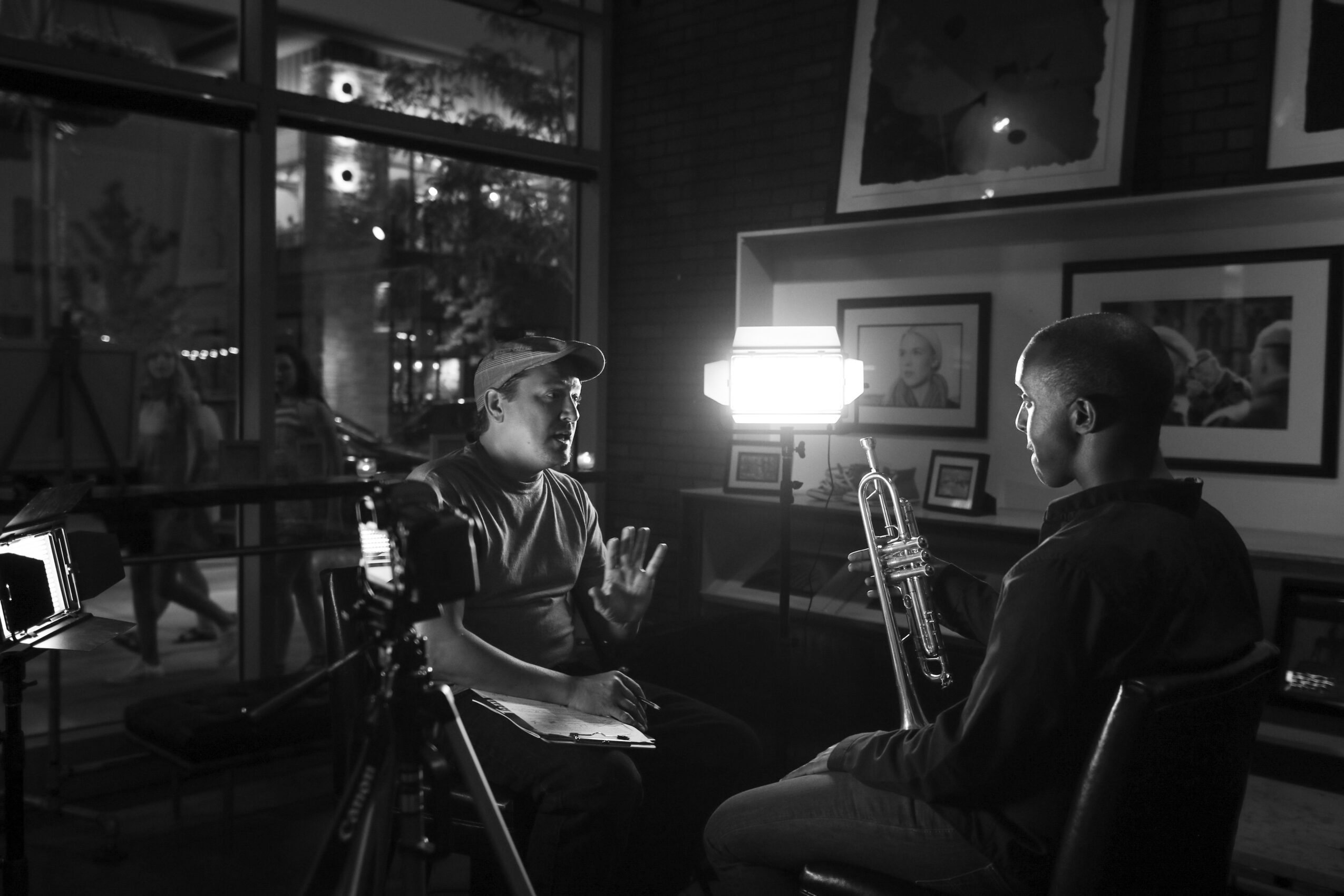 Two people sit and converse in a dimly lit room with framed pictures on the wall, one holding a saxophone, a camera nearby.