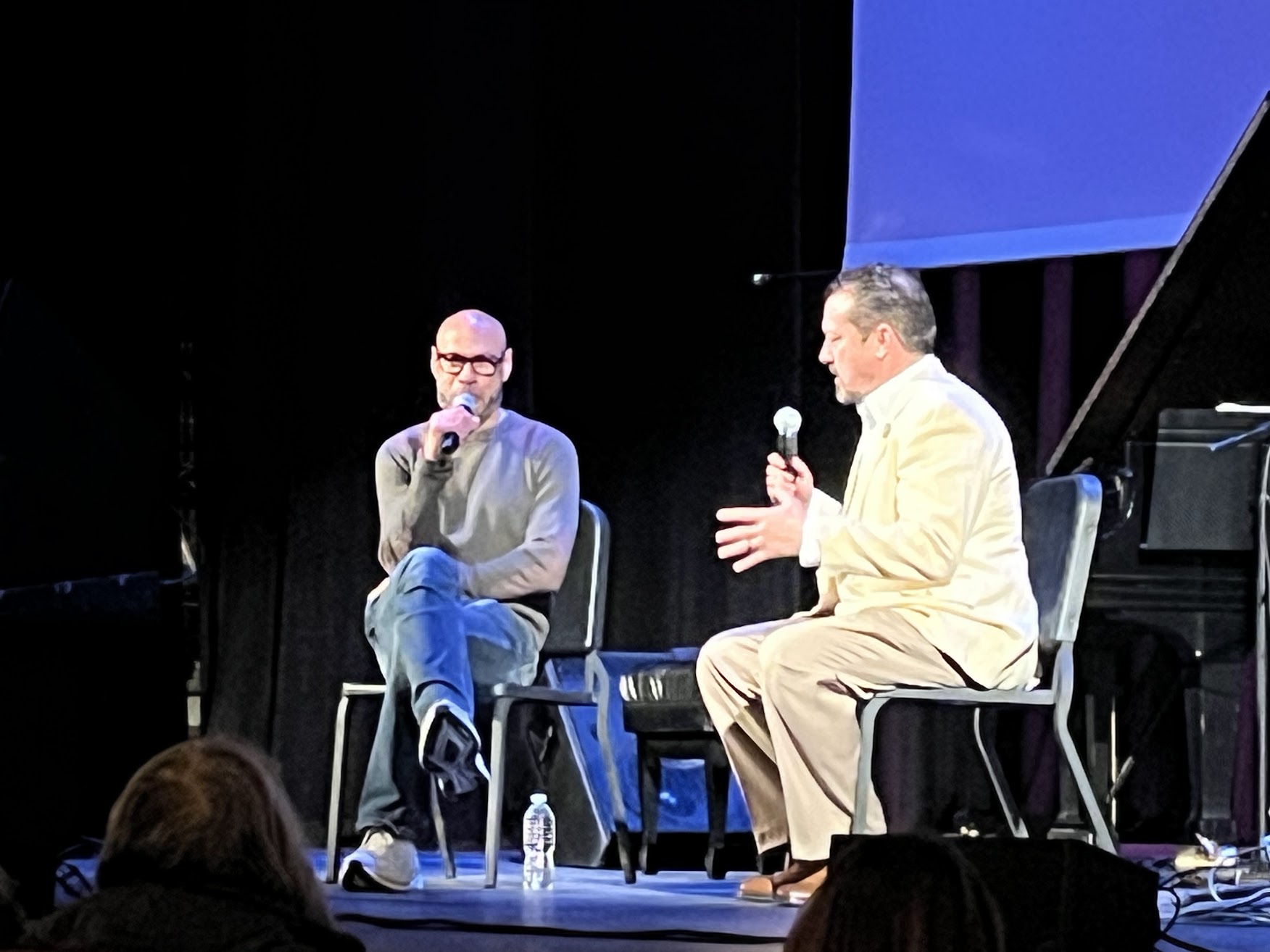 Two men sit on stage in conversation, one holding a microphone, with an audience visible in the foreground.