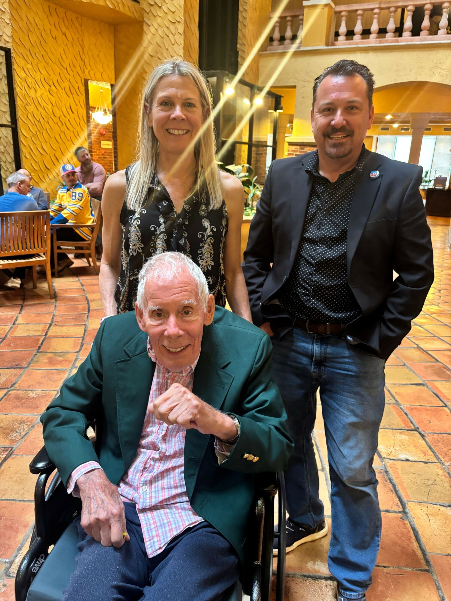 Three people, two men and one woman, pose together indoors, with brick flooring and warm lighting in the background.