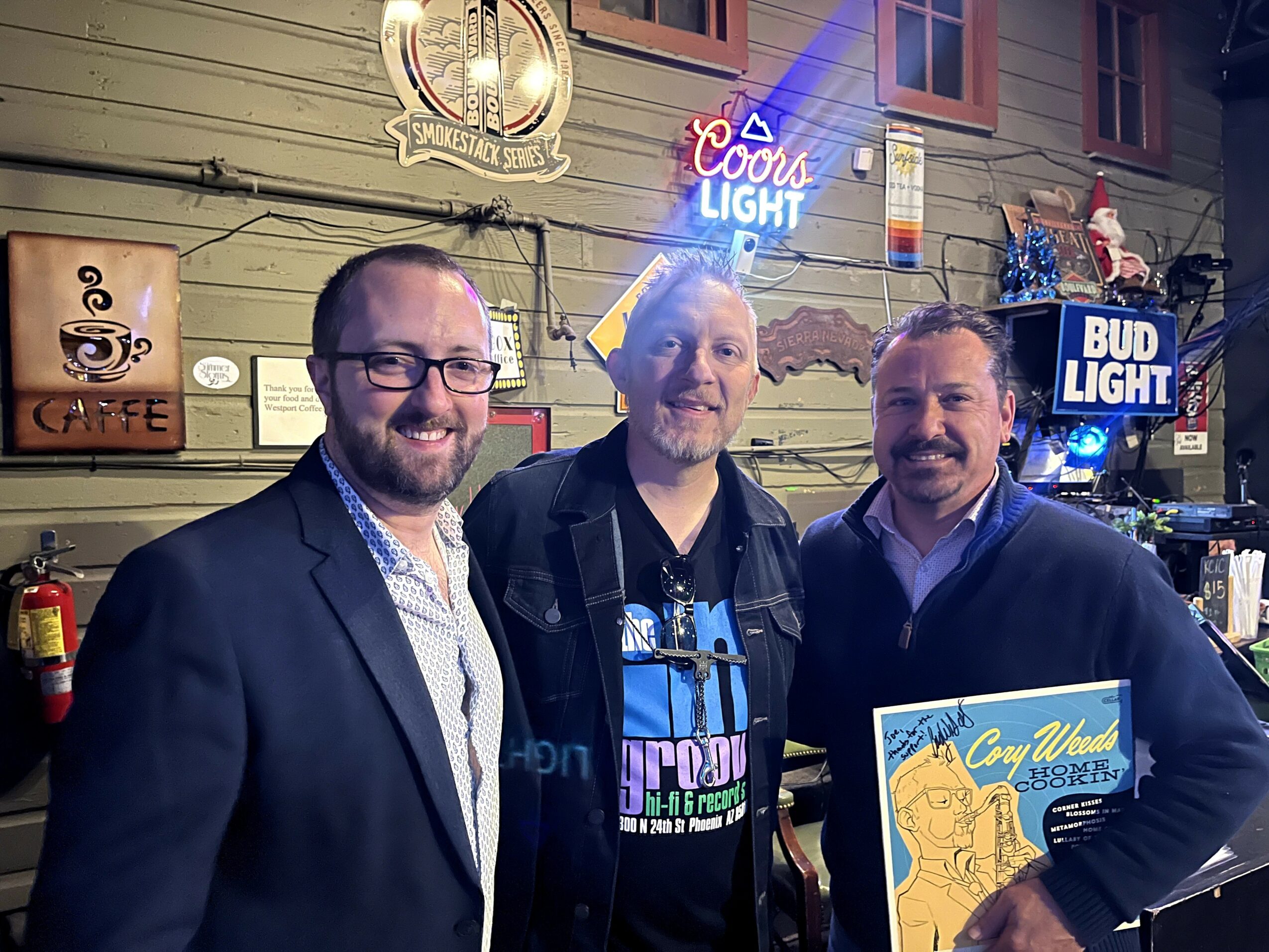 Three men standing outdoors at night, smiling, with neon signs and building in background.
