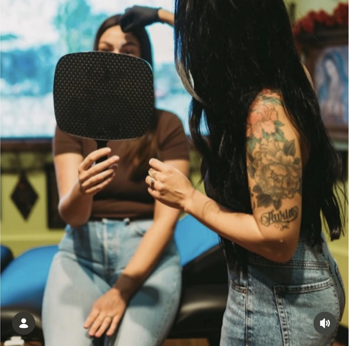 Two women with long dark hair, one with a tattoo, are in a room with a colorful background, one holding a mirror.