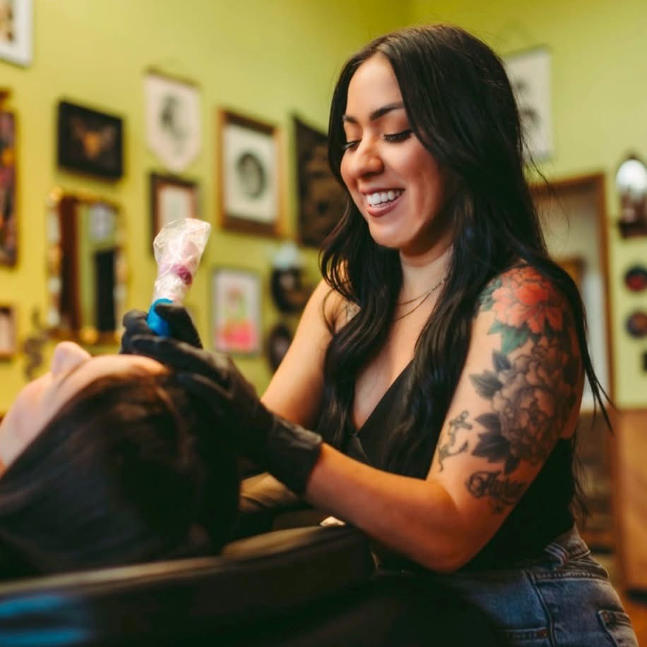 Woman with long dark hair and tattoos smiling while tattooing a person's arm in a colorful studio.