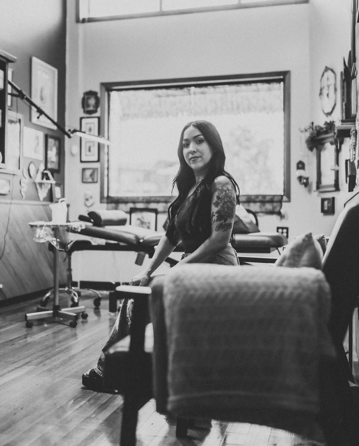 Woman with long hair sitting in a room with framed pictures on the wall and a large window behind her.