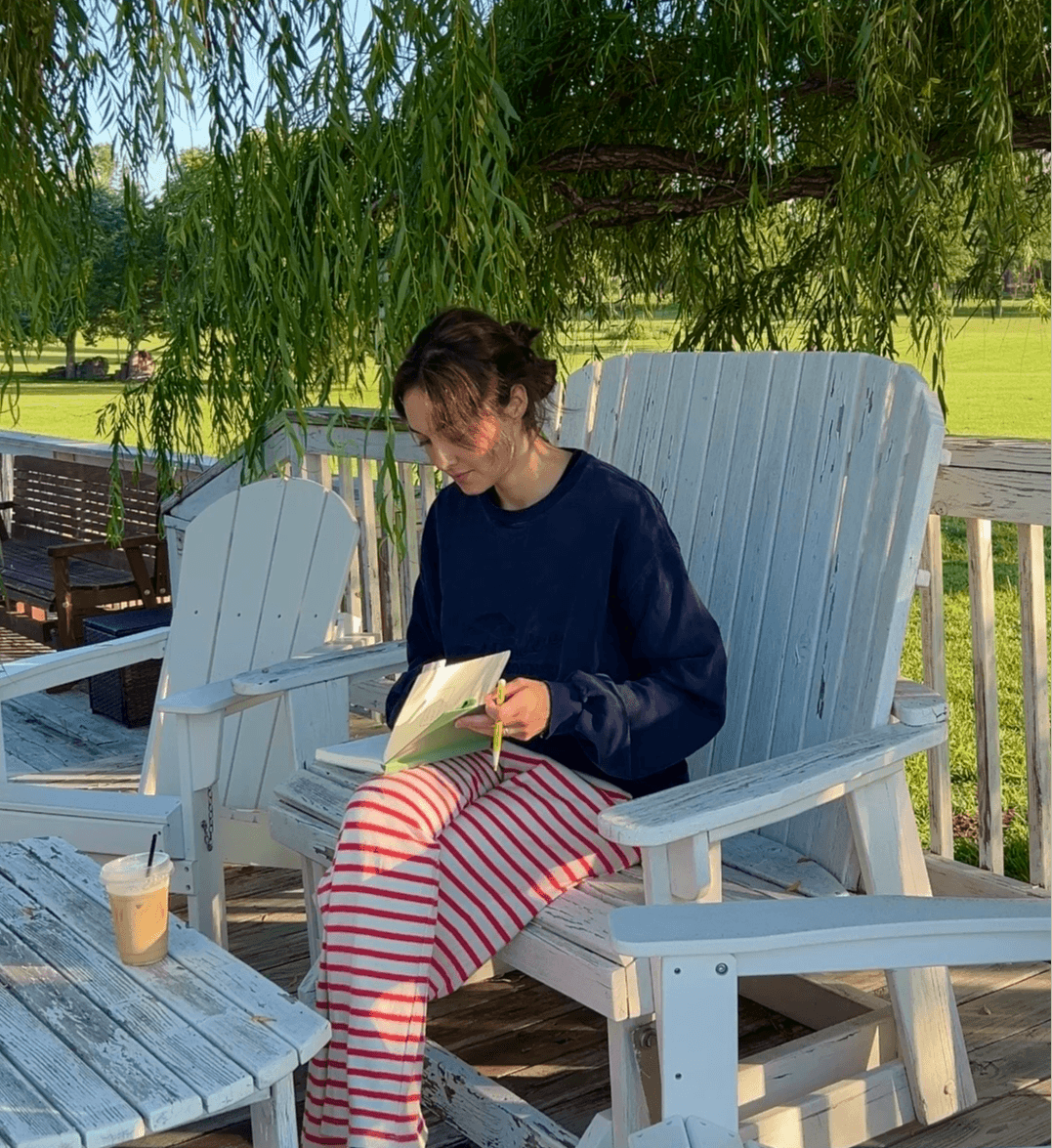 Person sitting on a white wooden chair reading a book outdoors under a tree, with a drink on a table nearby.
