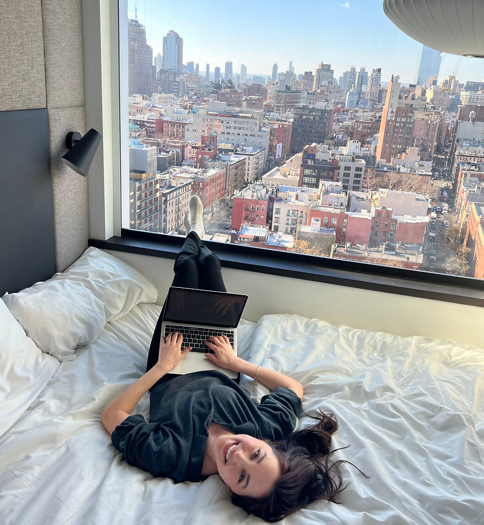 Woman lying on bed using laptop with city skyline view through large window behind her.