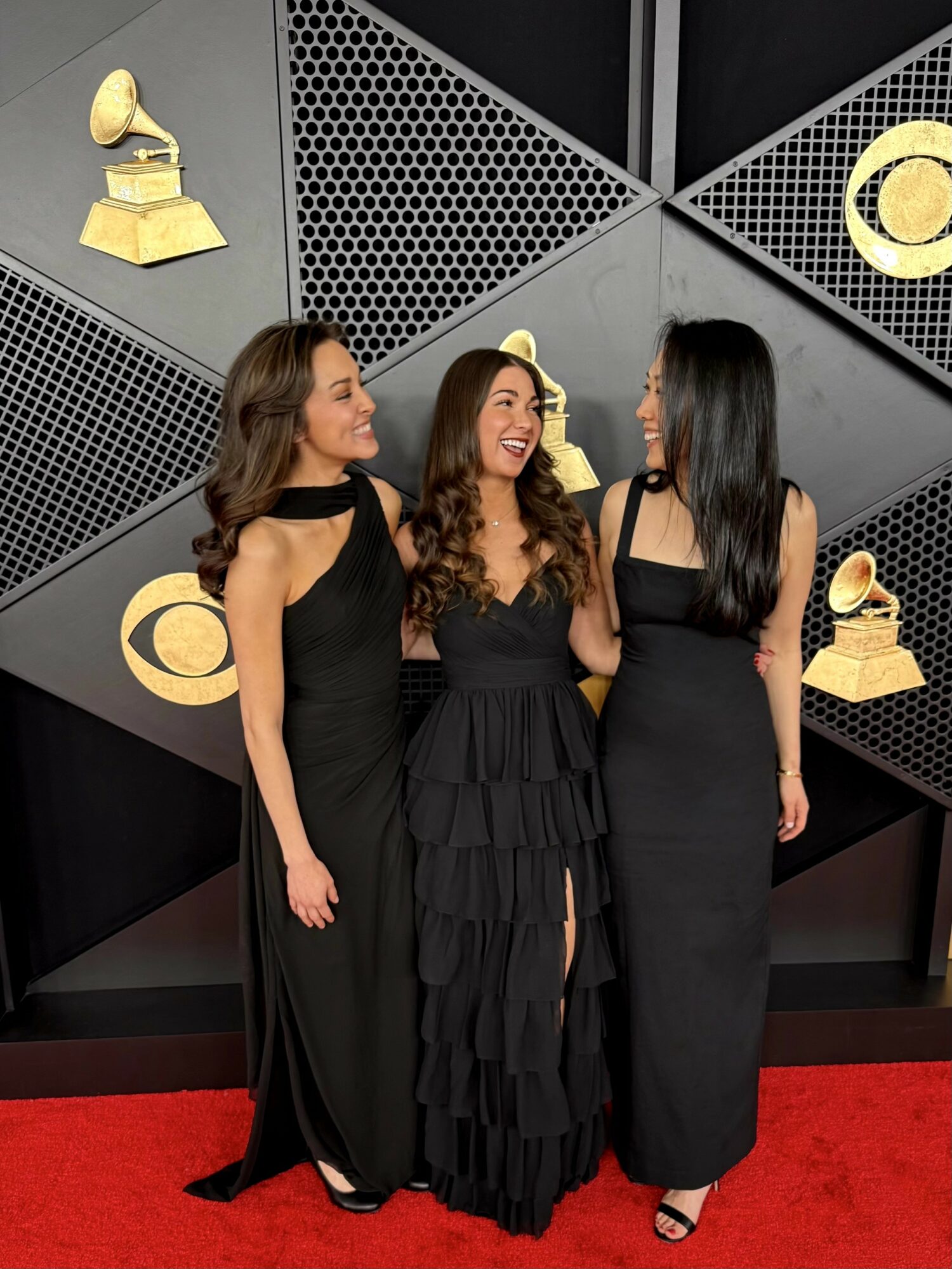Three women in black dresses standing on a red carpet, smiling and talking, with a geometric backdrop featuring Grammy awards.