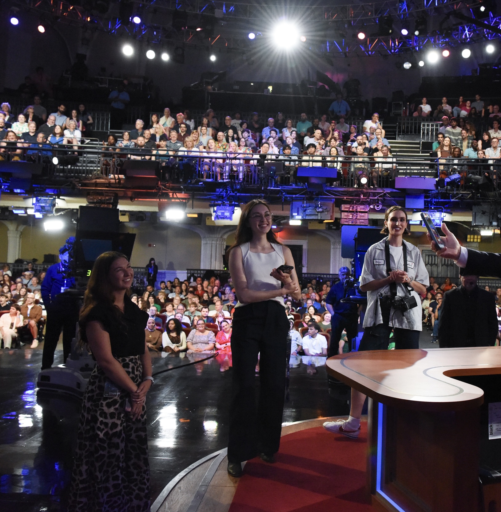 Three women stand on stage in front of a large audience, with bright lights overhead and a curved desk in the foreground.