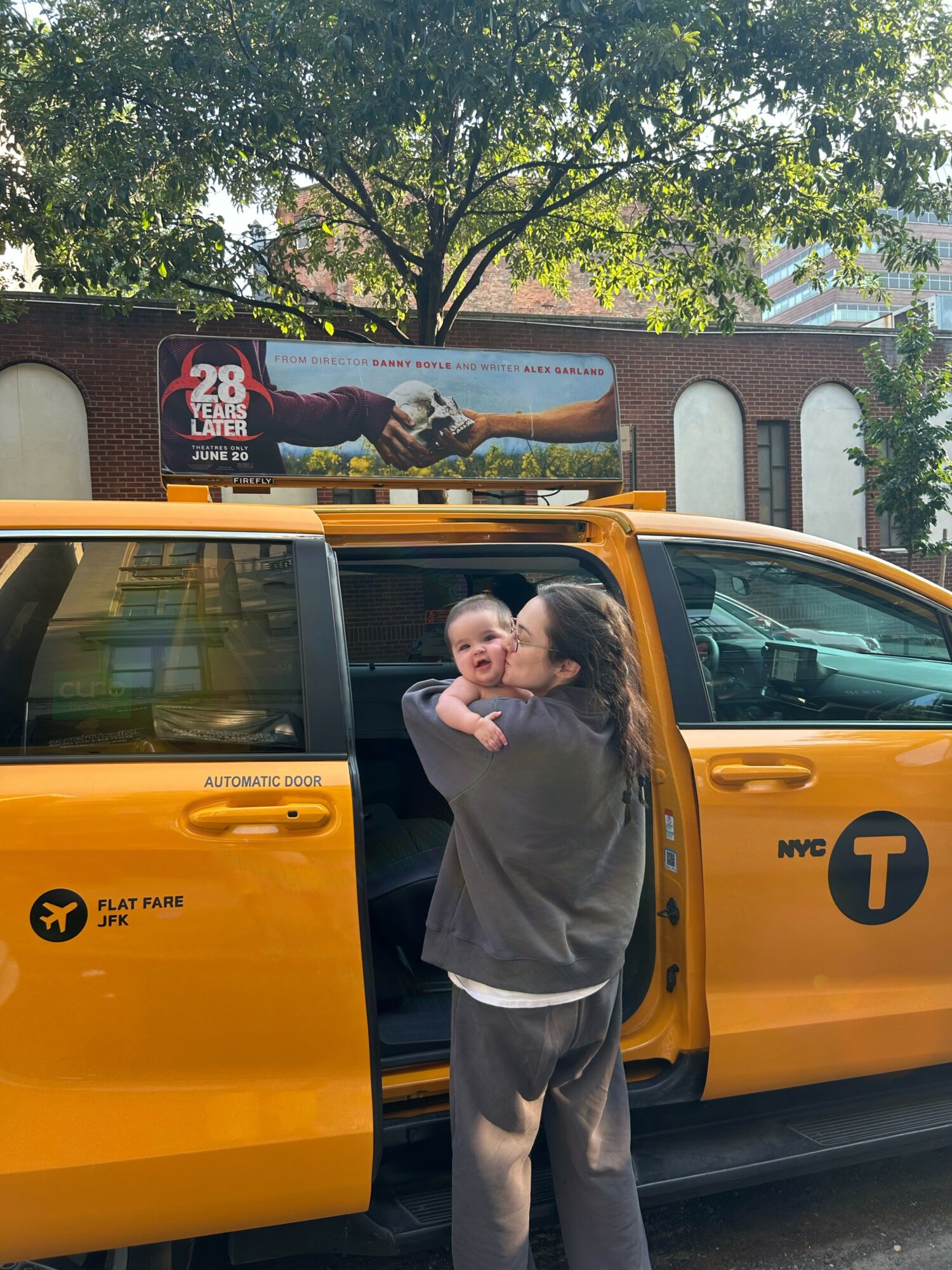 Person holding a baby next to a yellow NYC taxi cab, with a billboard and trees in the background.