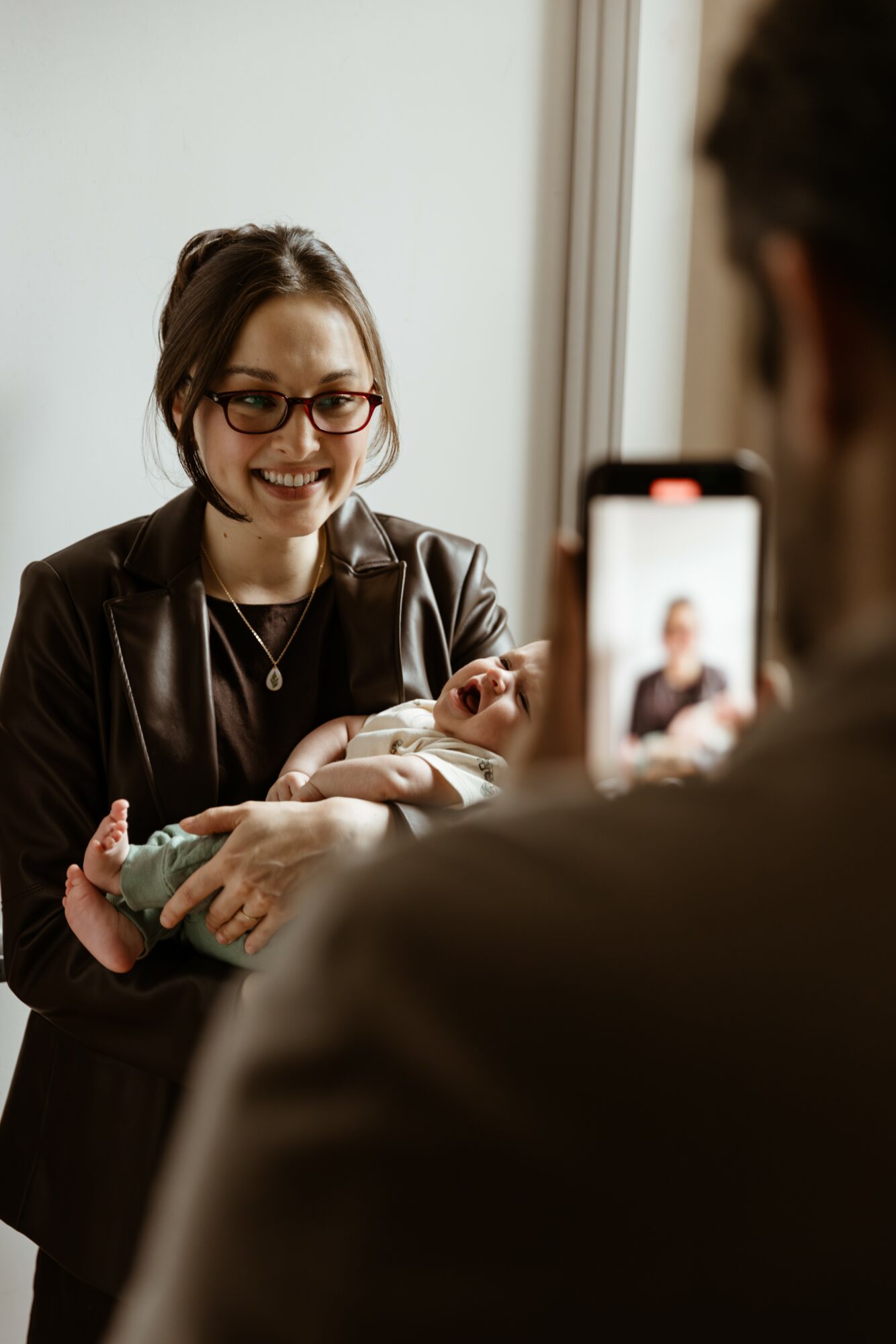 Woman smiling and holding a newborn baby, taking a photo with a smartphone, in a well-lit room.