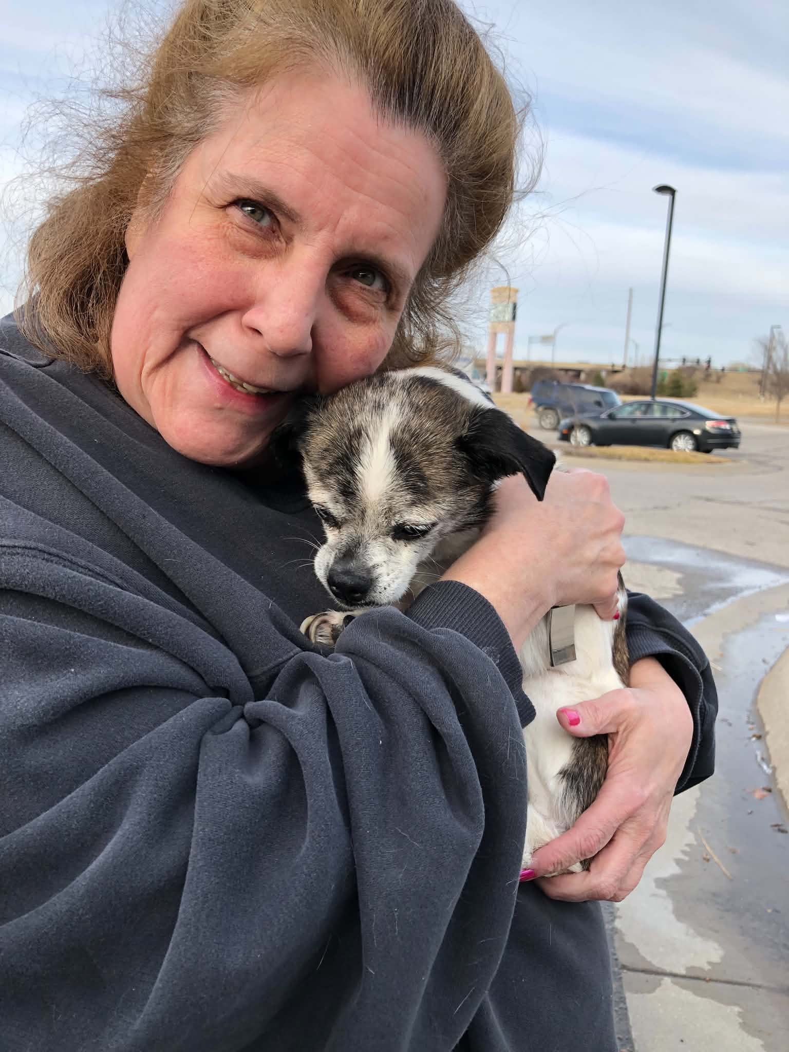 Woman holding a small puppy outdoors, smiling, with cars and a parking lot in the background.