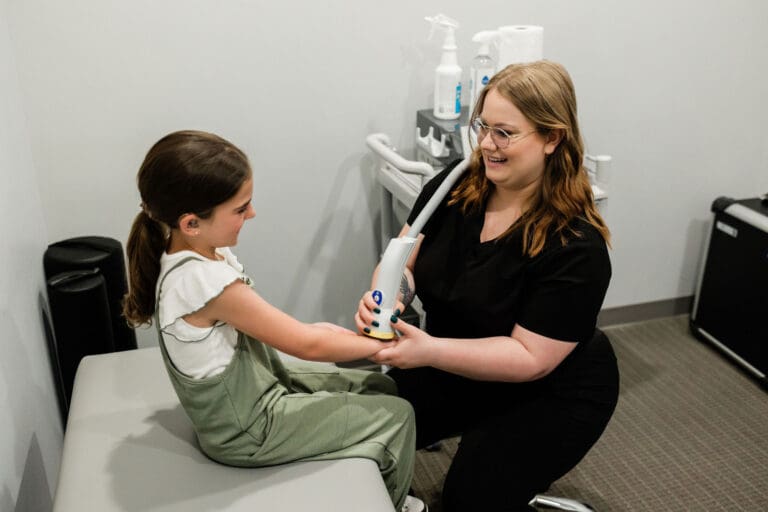 A young girl sitting on an examination table receives a medical device from a smiling healthcare worker in a clinical setting.