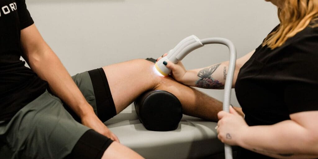Person receiving treatment with a device on their thigh, lying on a medical table, with another person assisting.