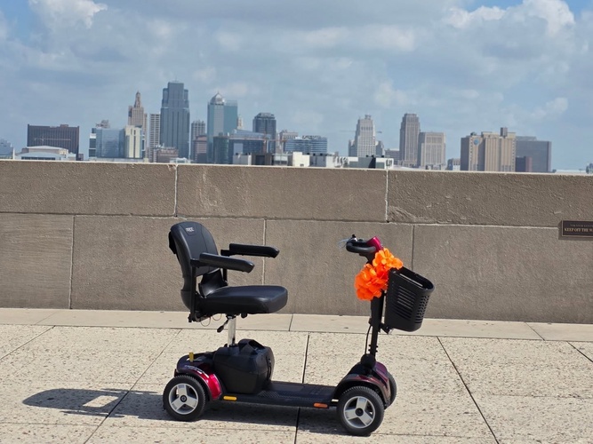 Electric scooter with a black seat and basket, parked on a sidewalk with a city skyline in the background.