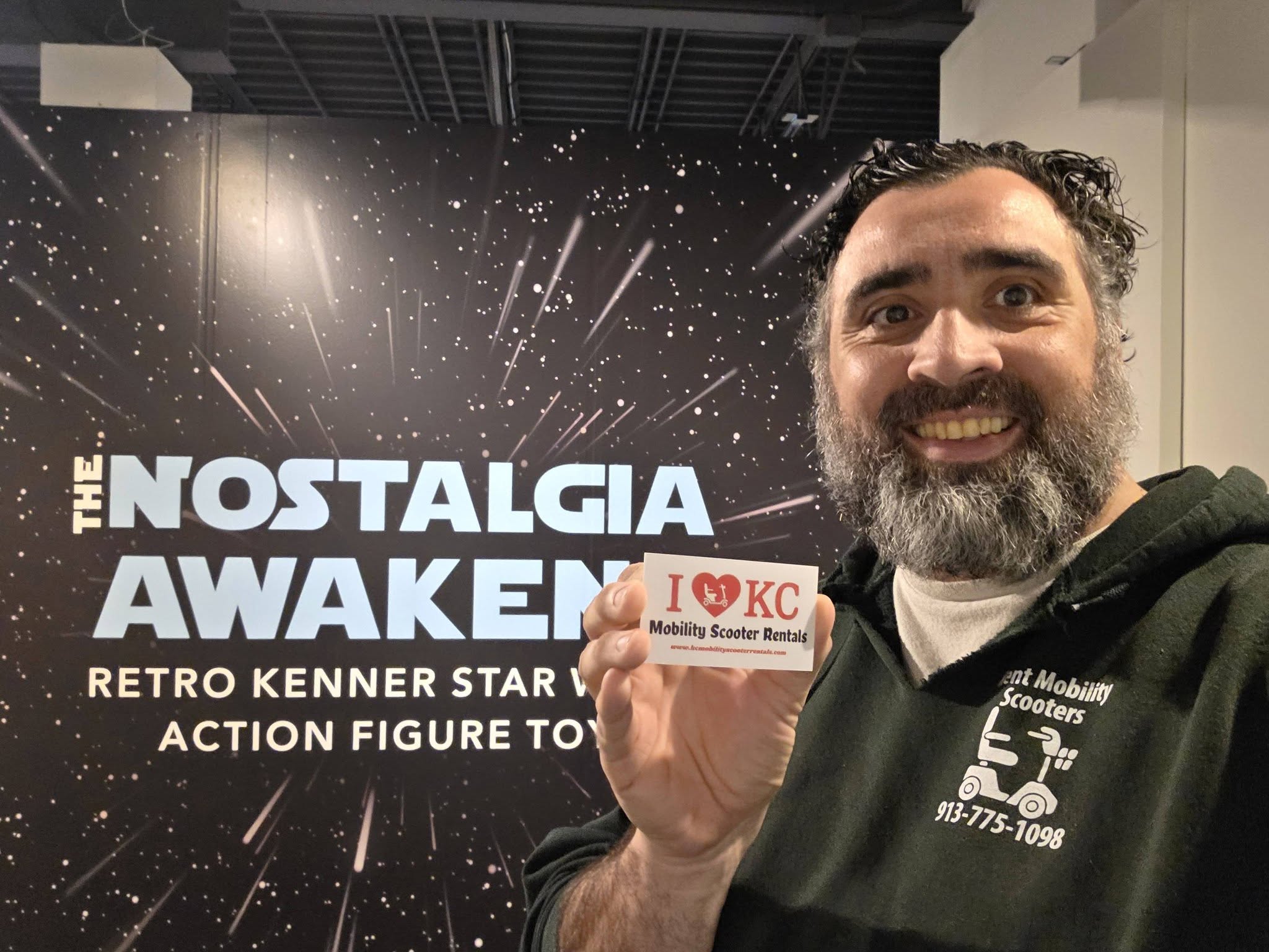 Man with a beard smiling holding a sign in front of a space-themed backdrop with text about nostalgia and action figures.