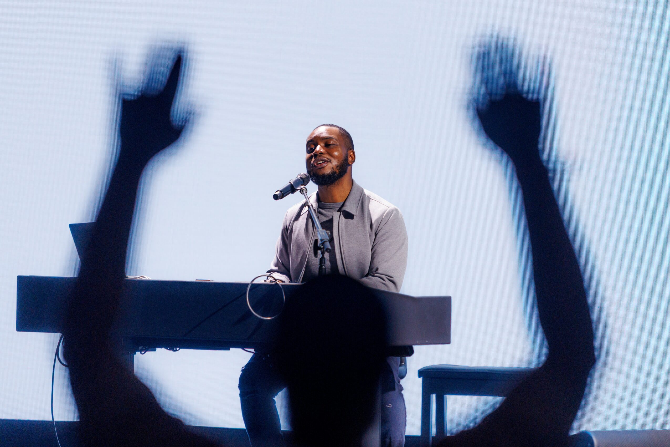 Man speaking at a podium with raised hands in the foreground, blurred audience, and a light background.