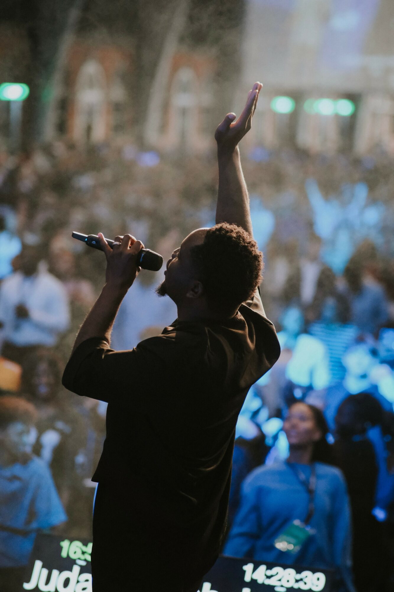 Silhouette of a person singing into a microphone with one arm raised, performing on stage with a crowd in the background.
