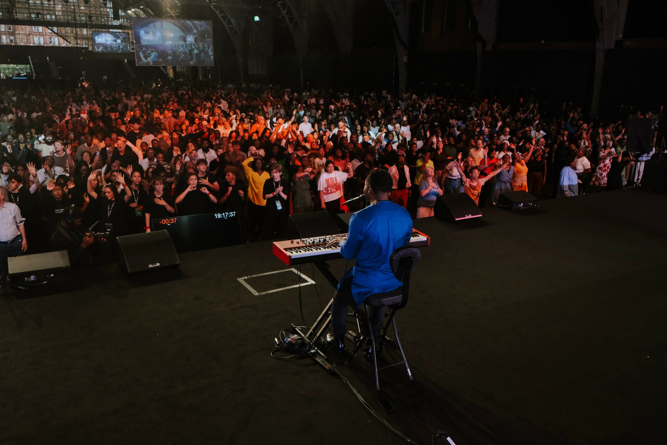 Performer in blue shirt on stage with a keyboard facing a large audience in a dark venue.