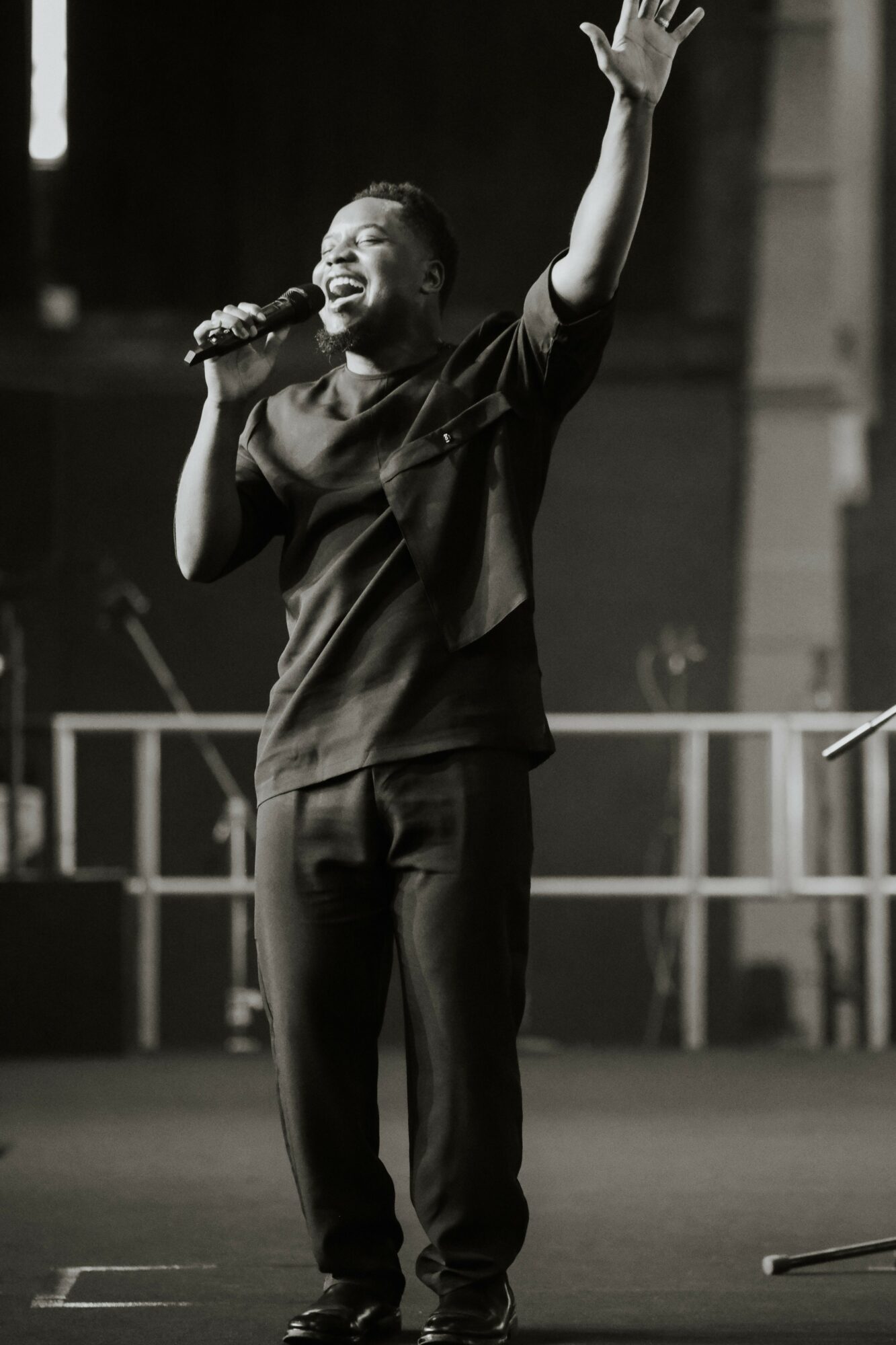 Person singing on stage with one arm raised, holding a microphone, smiling, in a dark setting with stage equipment.