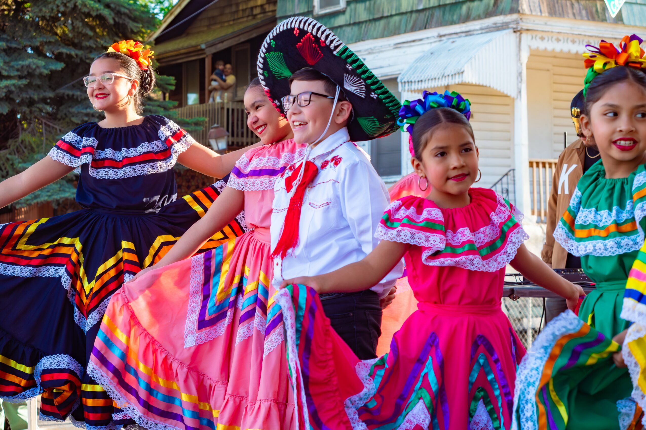 Group of children in colorful traditional dresses and sombrero, smiling and dancing outdoors.