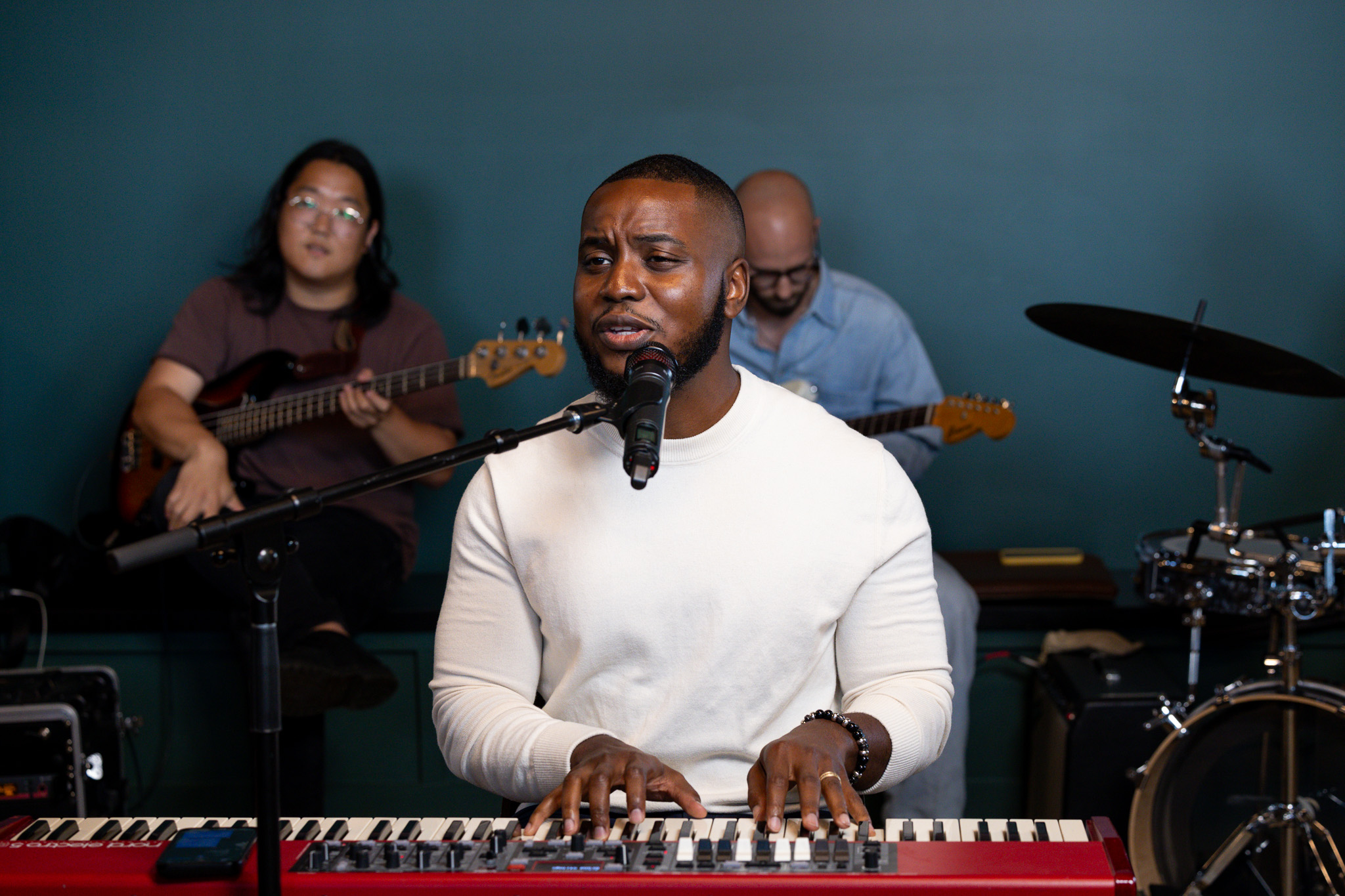 Three musicians perform with a man playing keyboard in front, a woman with guitar, and a man with drums in background.