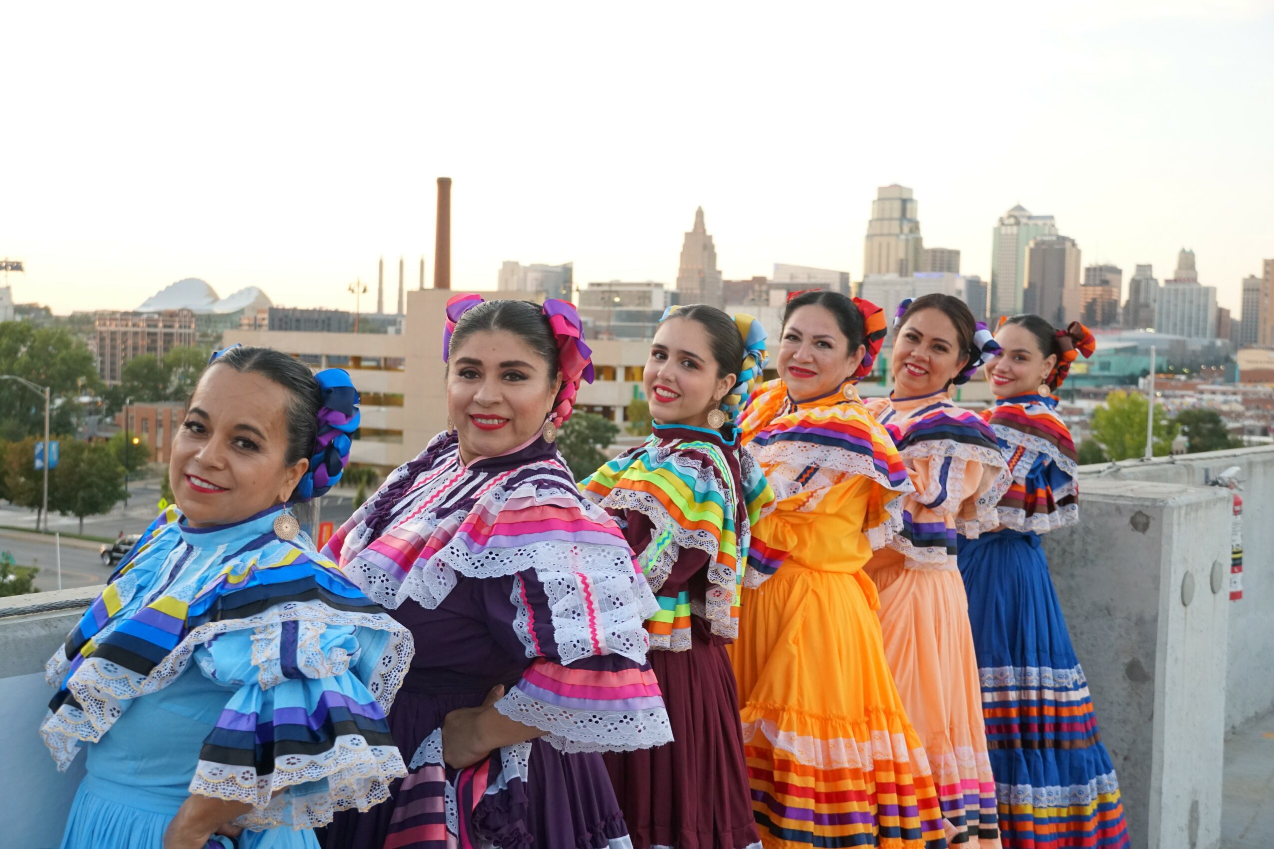 Six women in colorful traditional dresses standing on a rooftop with city skyline in background.