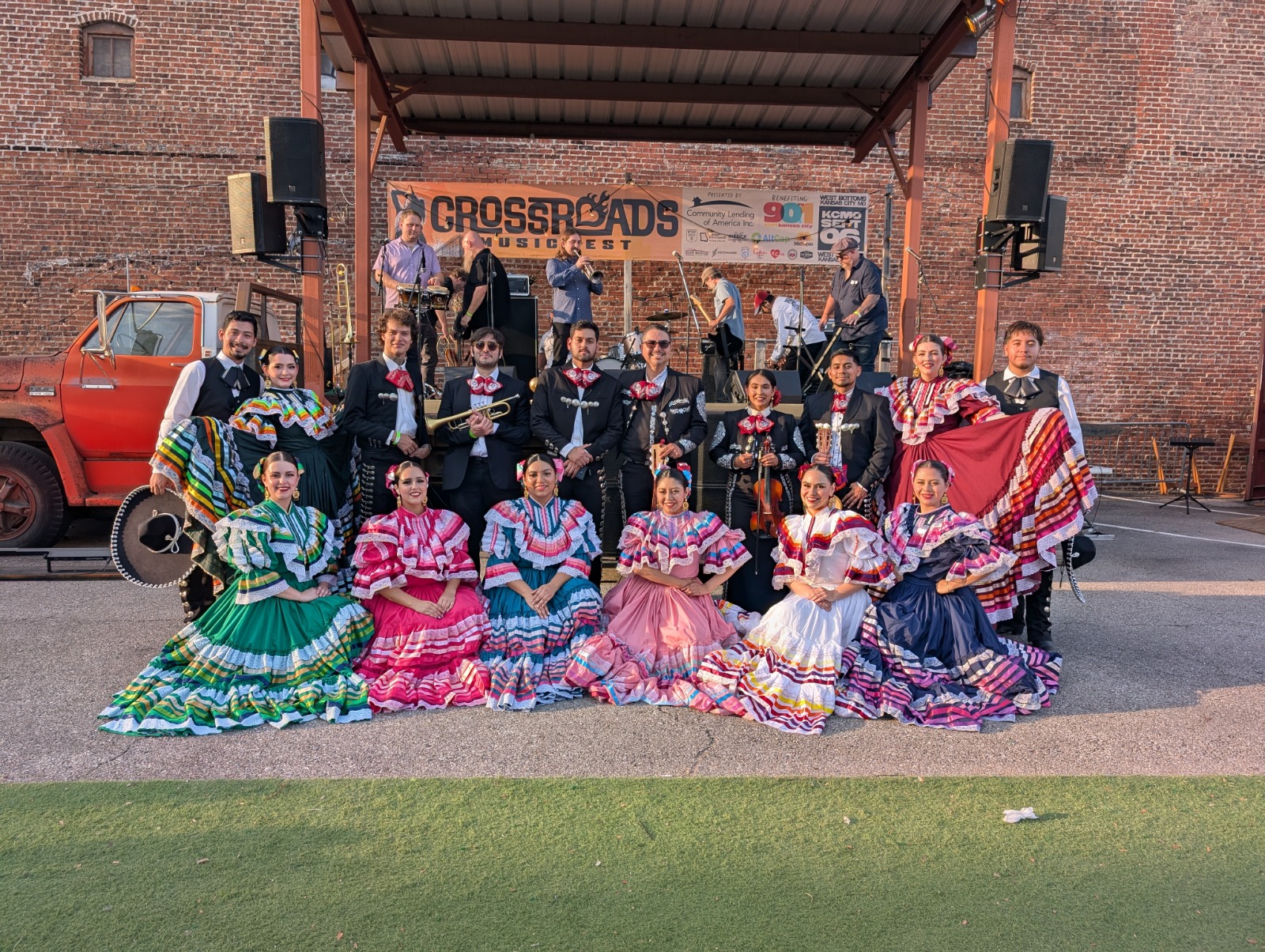Group of people in colorful traditional dresses posing outdoors in front of a stage with a banner, some holding shawls.