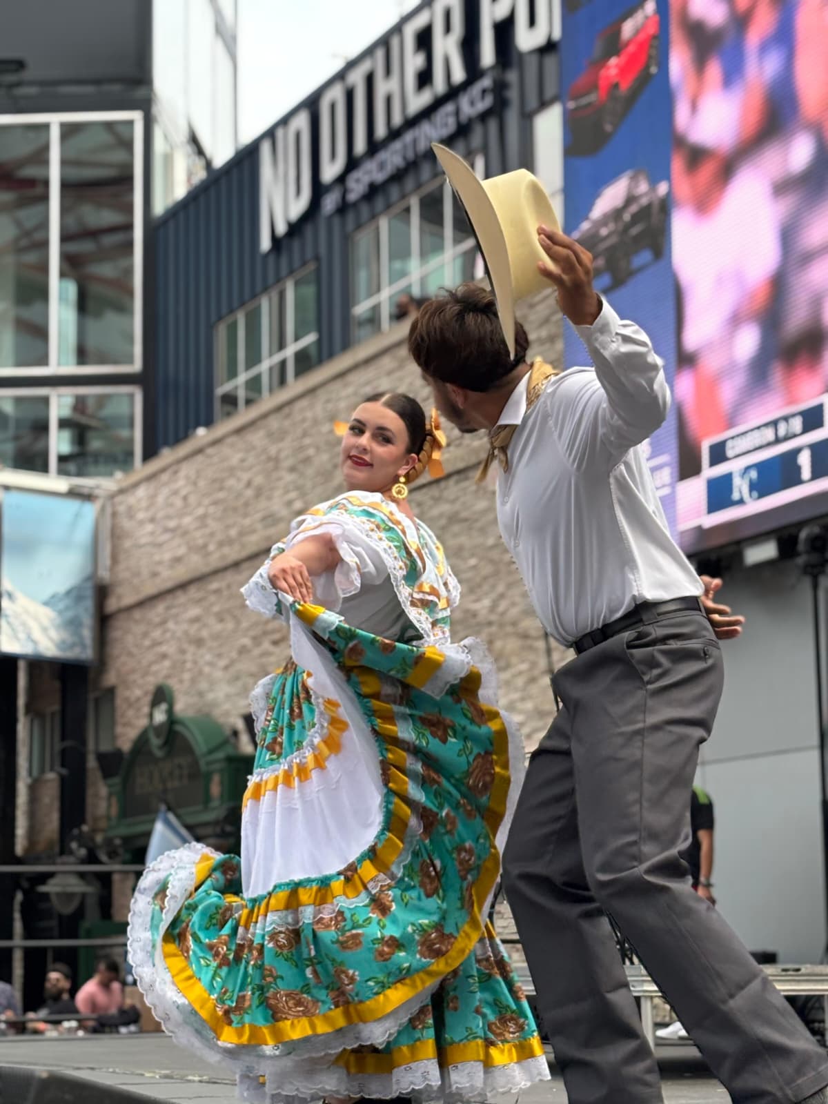 A woman in colorful traditional dress dances with a man in a white shirt and gray pants holding a hat, outdoors.