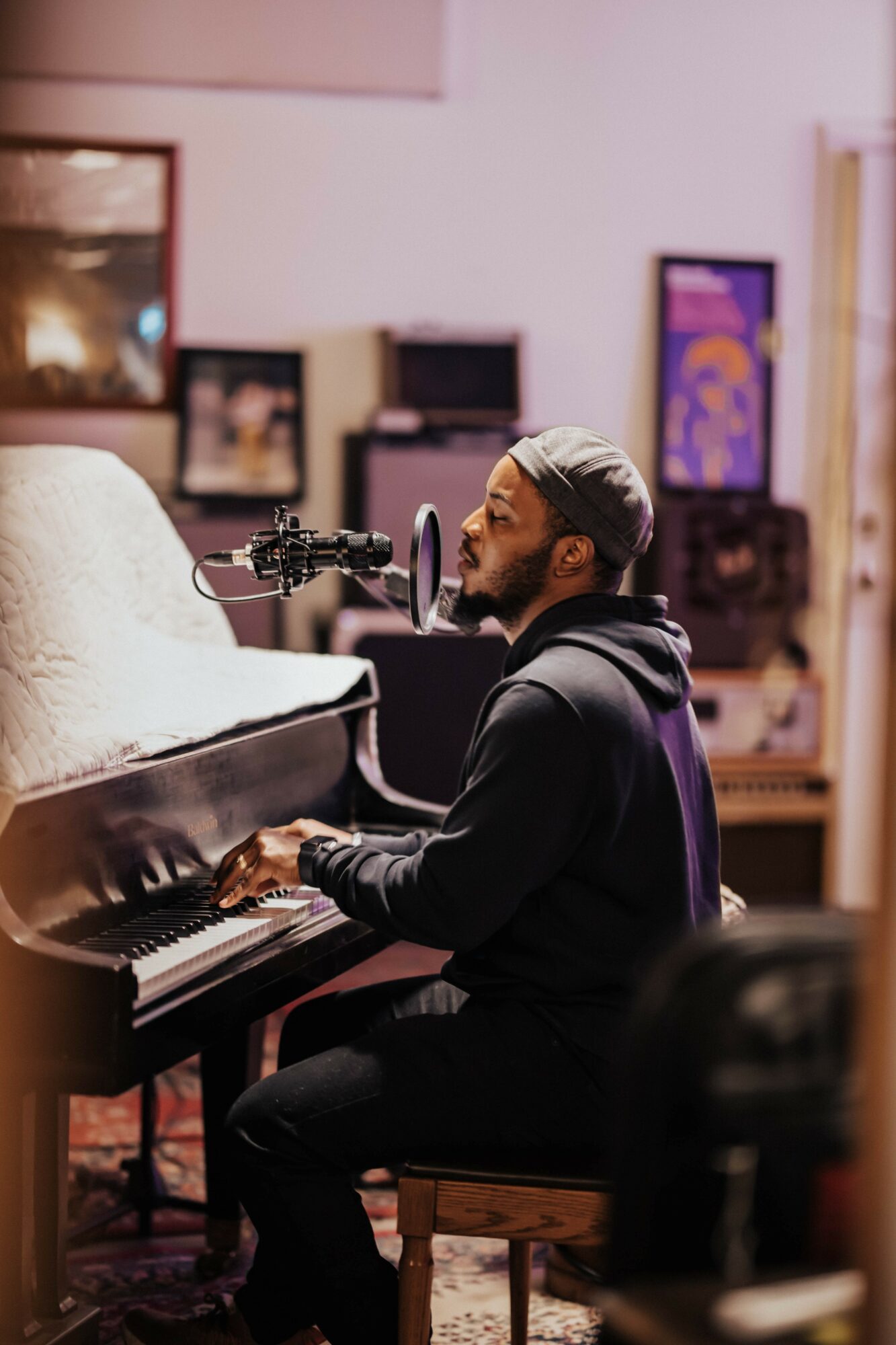 Man playing piano in a music studio with a microphone, surrounded by framed pictures and equipment.