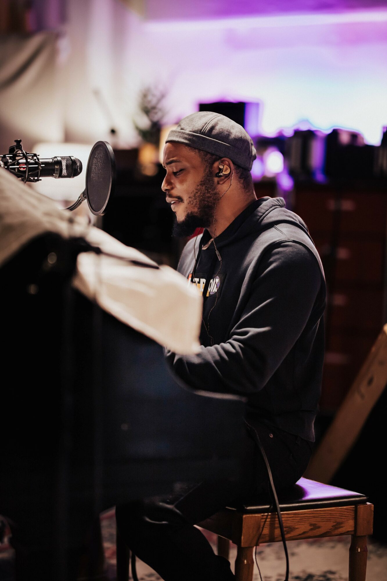 Person with a beard and gray beanie playing piano in a dimly lit room with purple lighting.