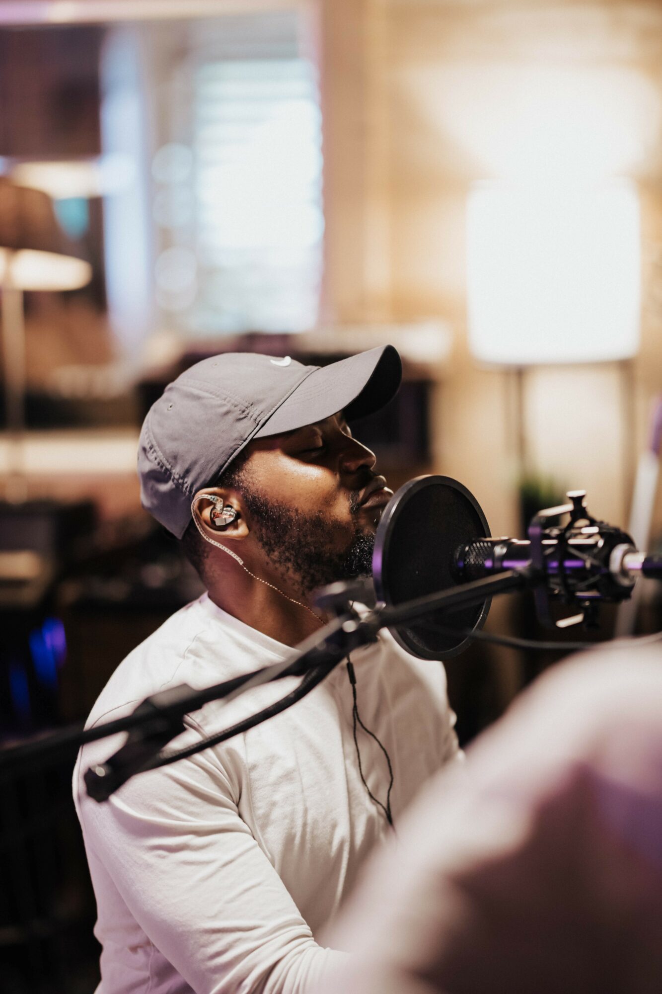 Man wearing a cap and glasses singing into a microphone in a recording studio, with blurred background.